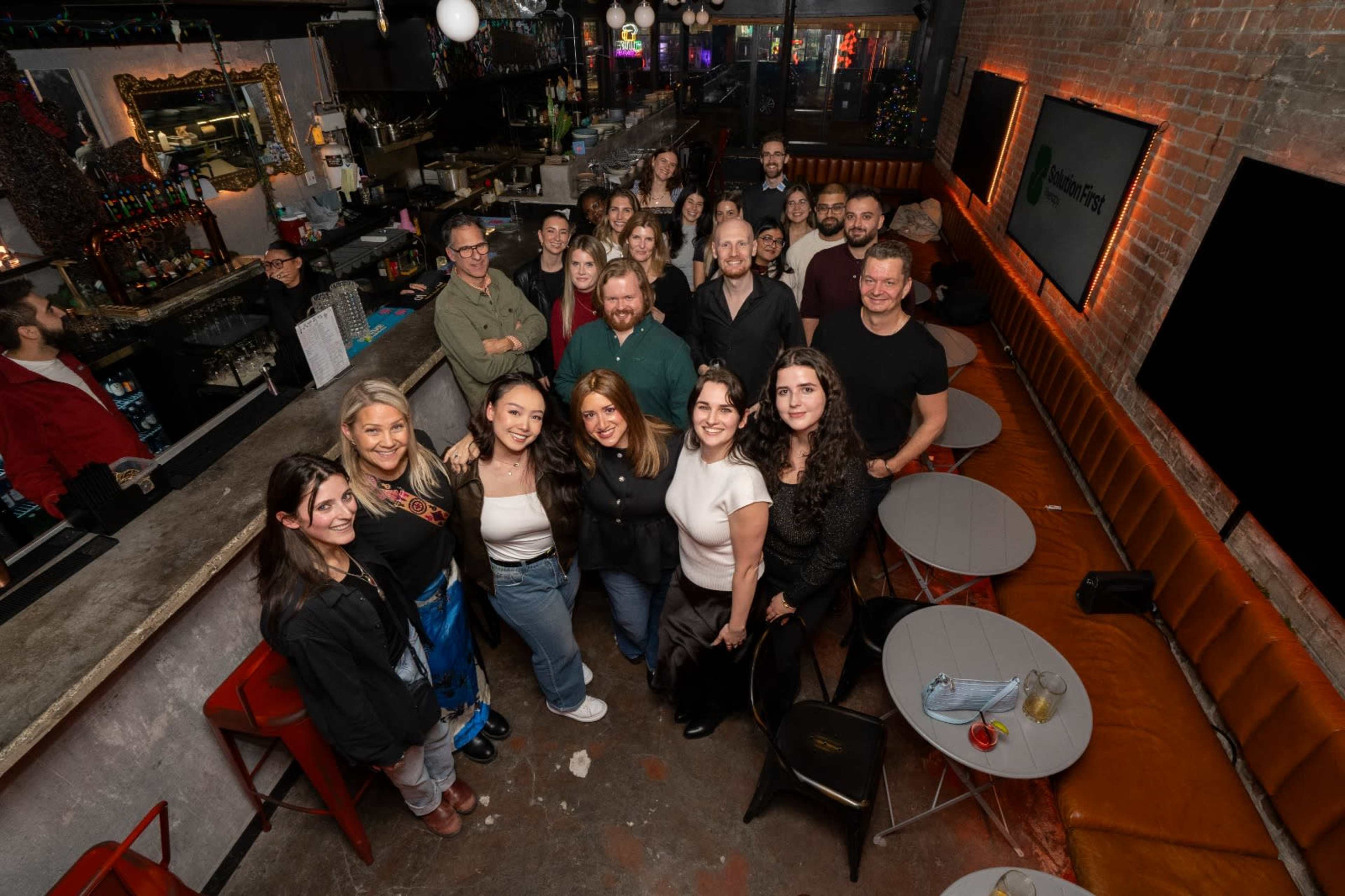 A diverse group of people gathers for a photo in a lively bar setting with soft lighting and a brick wall backdrop.