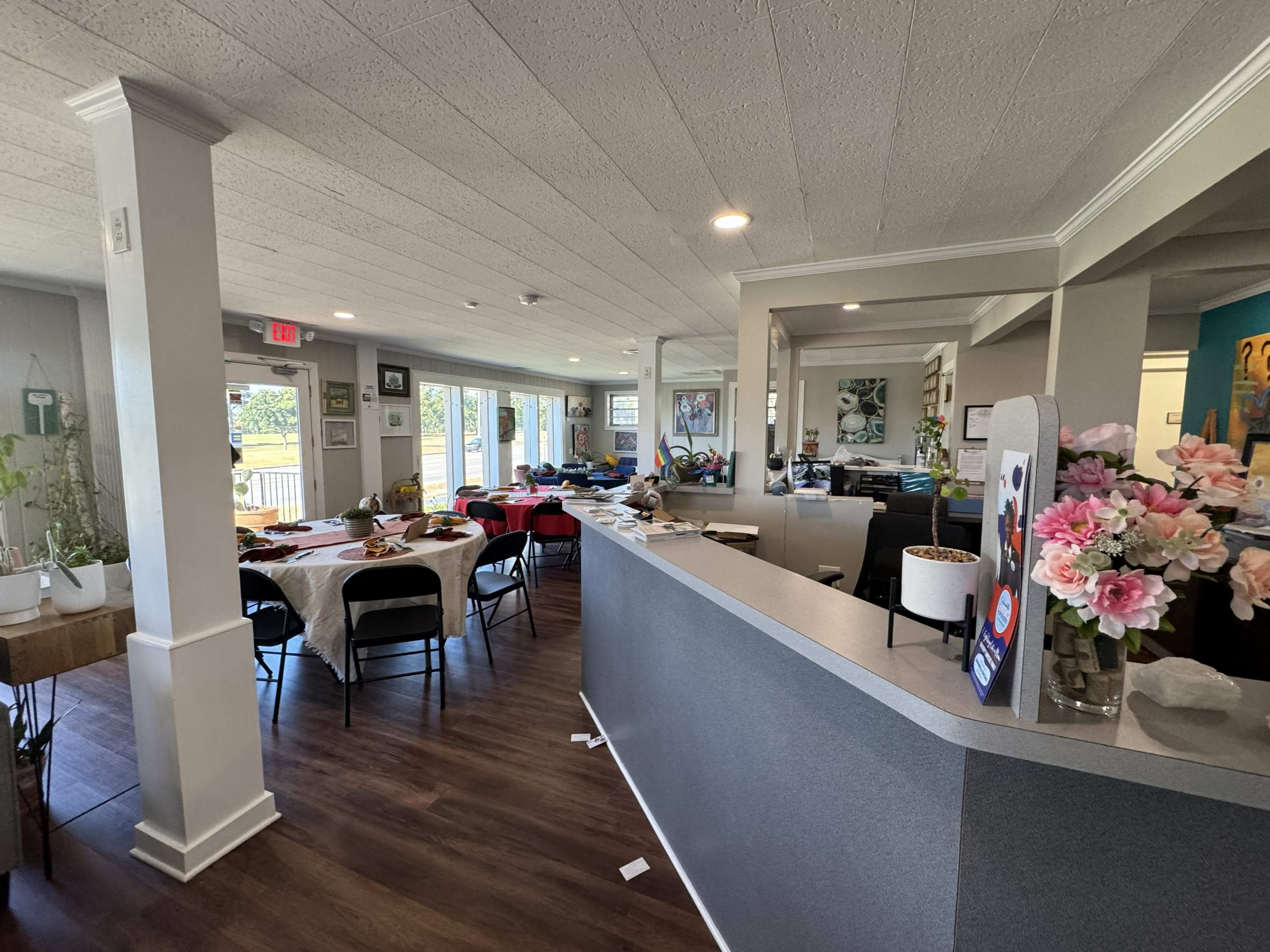 The image shows a bright interior of a restaurant or café with multiple tables set for dining, a reception area, and a mix of decorative elements on the walls.