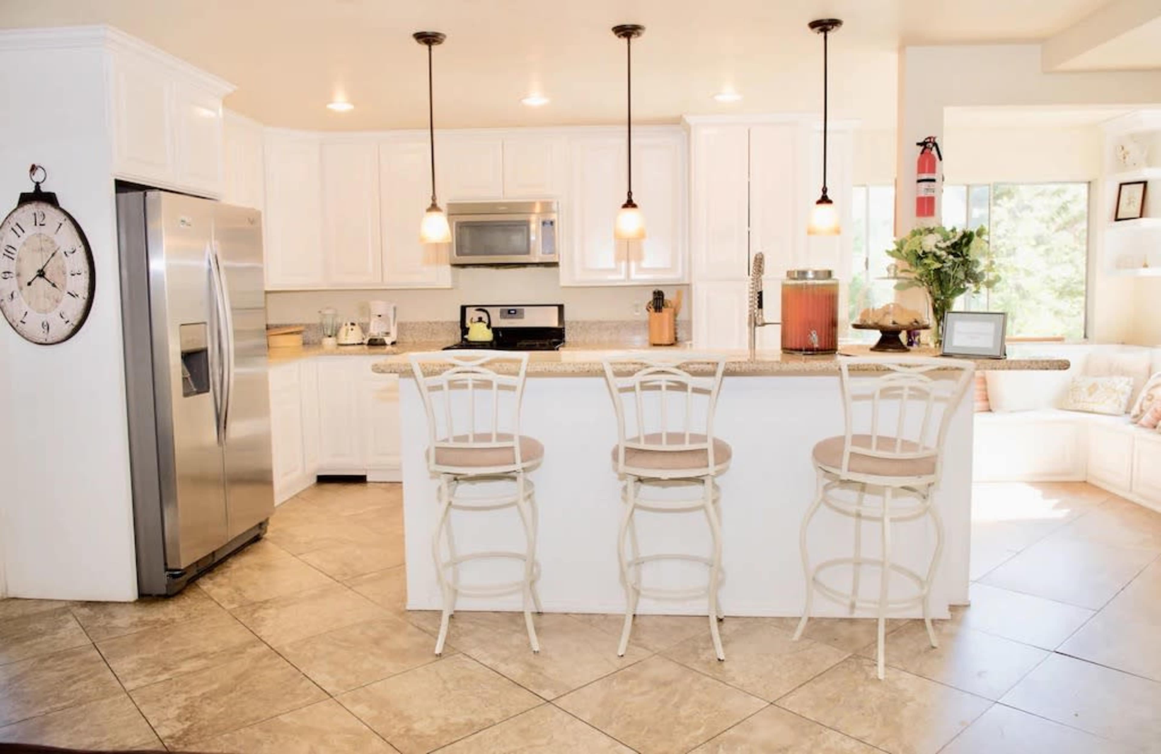 The image shows a modern kitchen with white cabinetry, stainless steel appliances, and three bar stools at a countertop island.