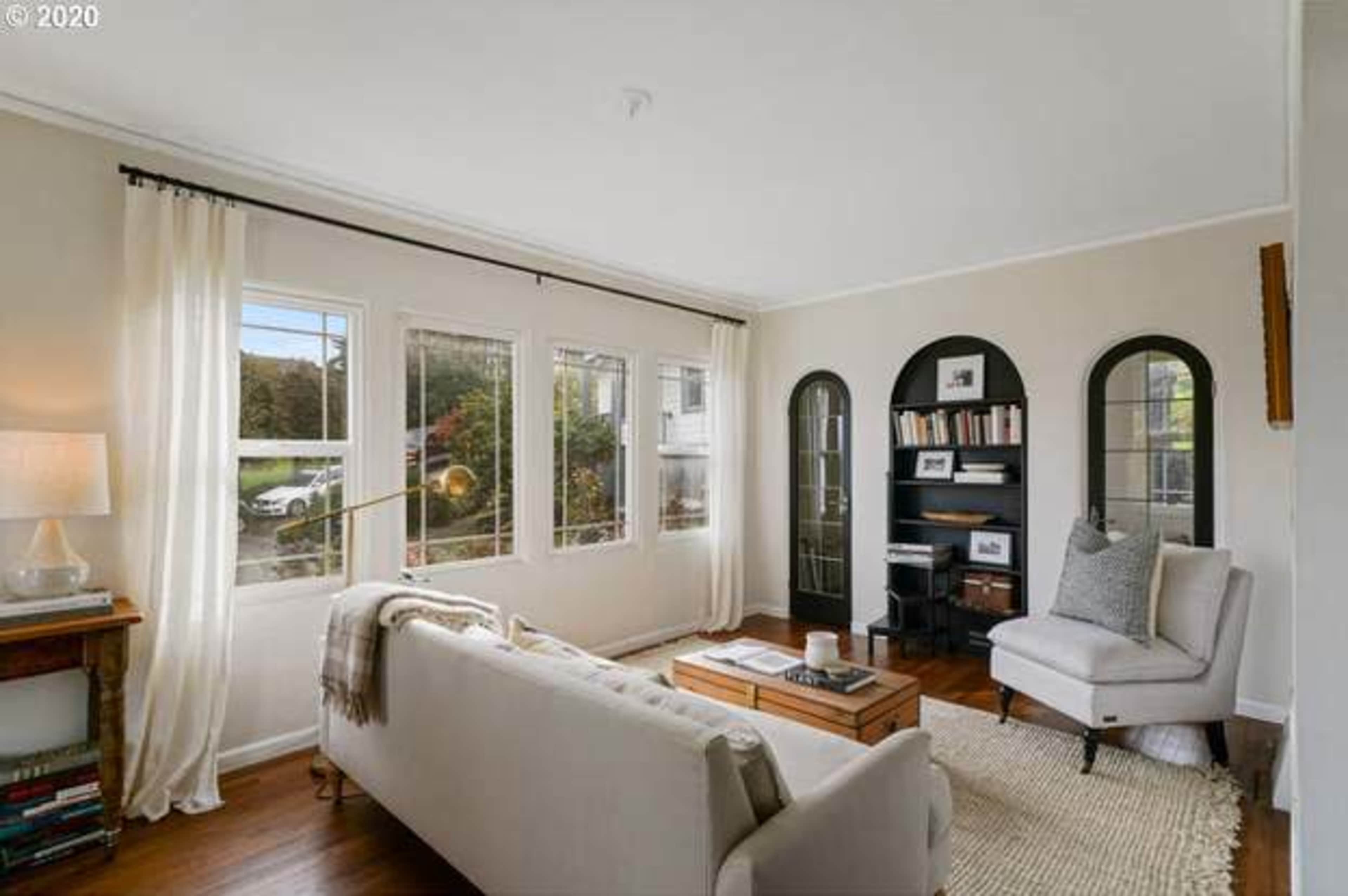 A cozy living room features a white sofa, a wooden coffee table, and large windows allowing natural light, with bookshelves and decorative arches in the background.