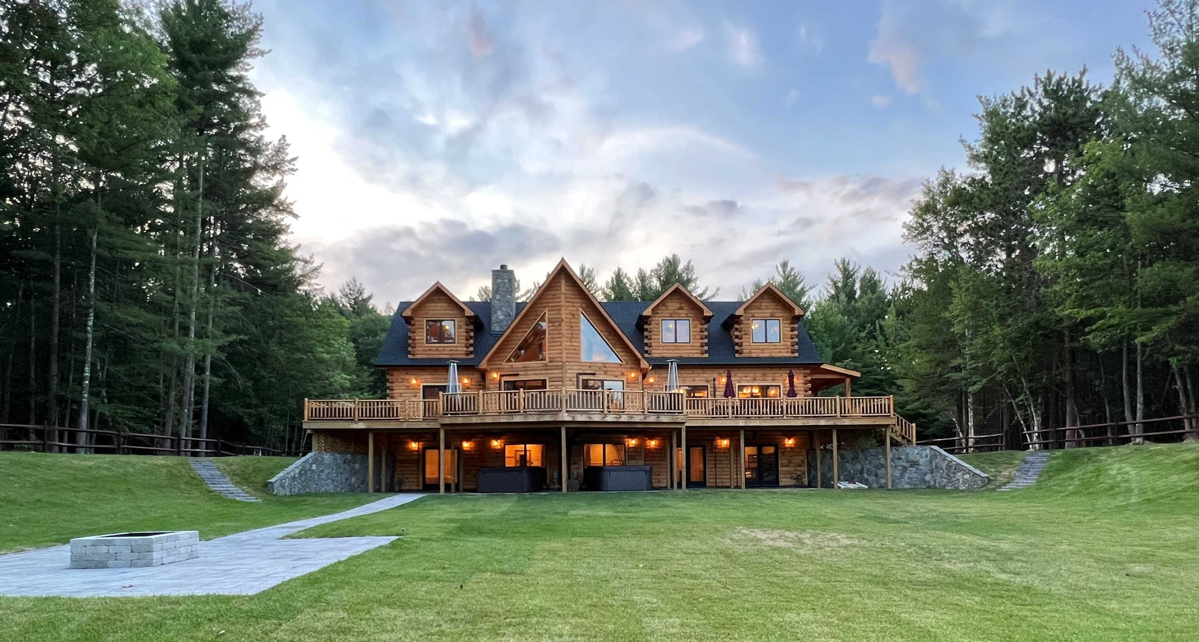 A large wooden house with multiple gabled roofs is situated on a grassy lawn surrounded by trees, featuring a spacious deck and stone landscaping.