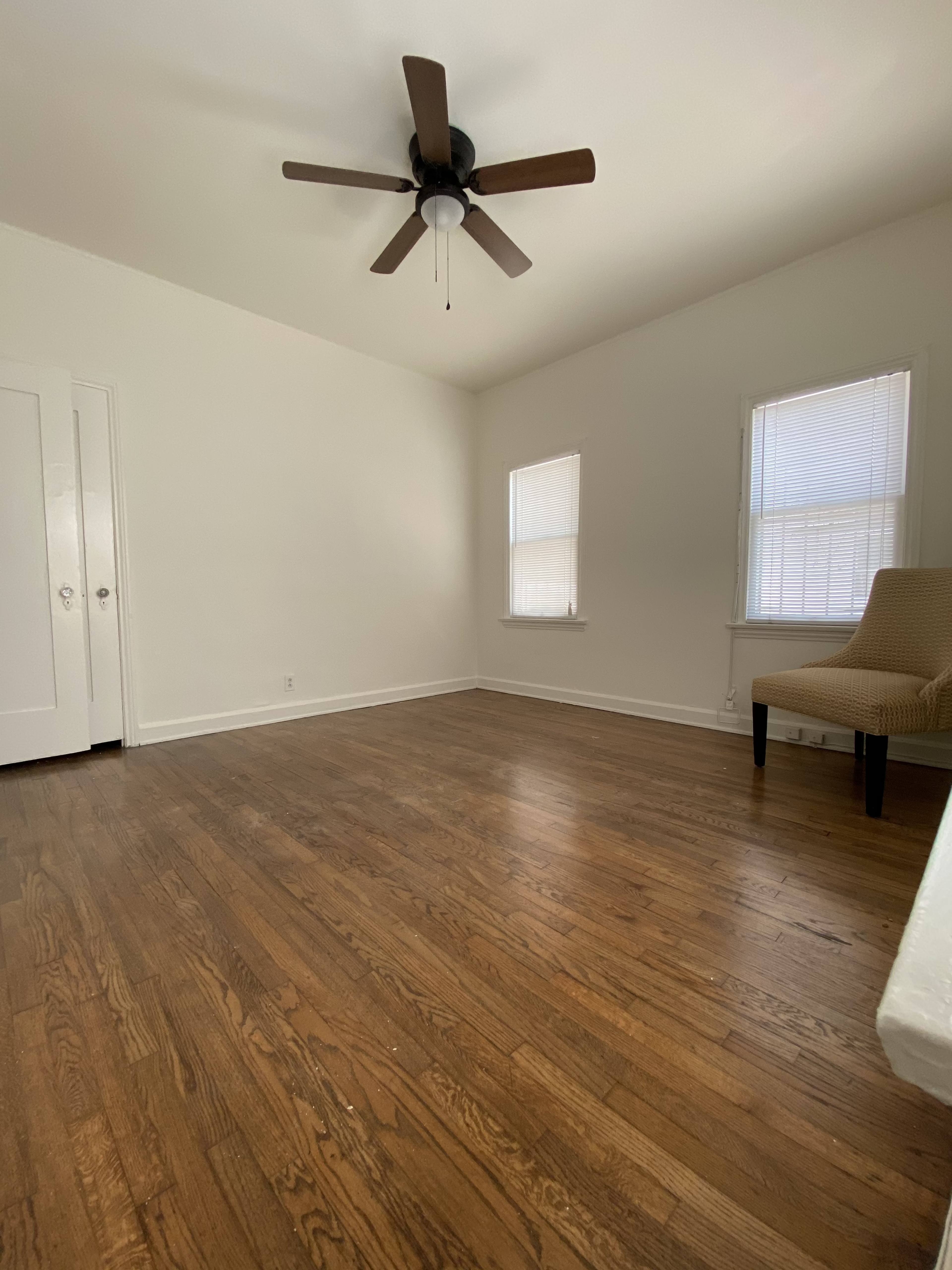 The image shows a spacious, empty room with hardwood flooring, a ceiling fan, and two windows.