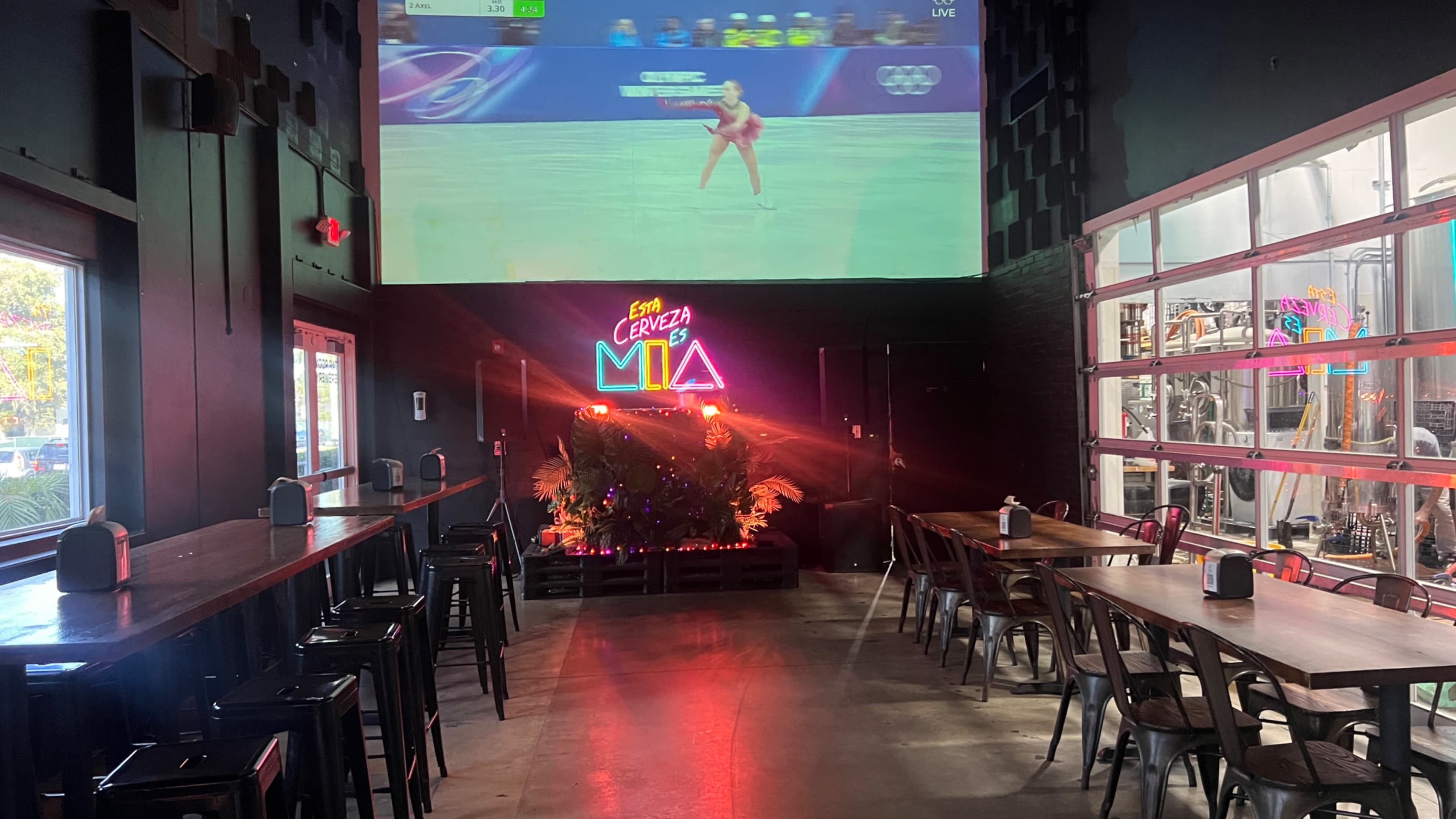 The image shows a dimly lit indoor space with long tables and metal chairs, featuring a large screen displaying a live sporting event and a colorful neon sign that reads "Cerveza Mia."