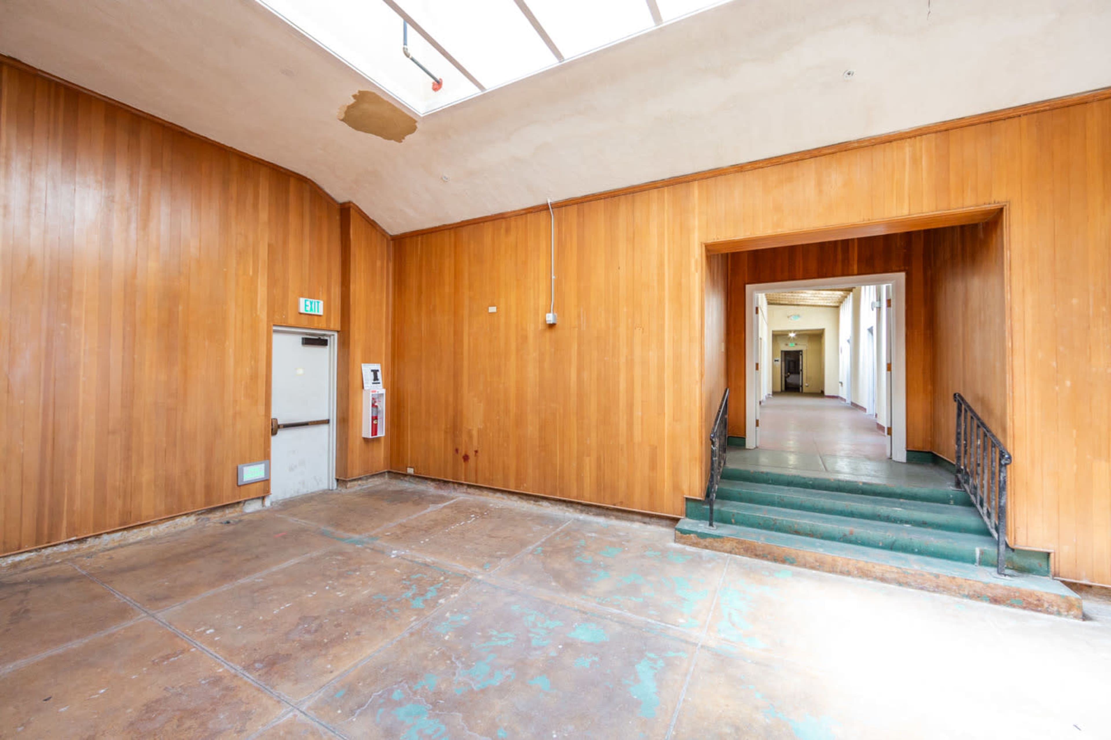 The image shows an empty room with wooden paneling and a doorway leading to a staircase in the background.