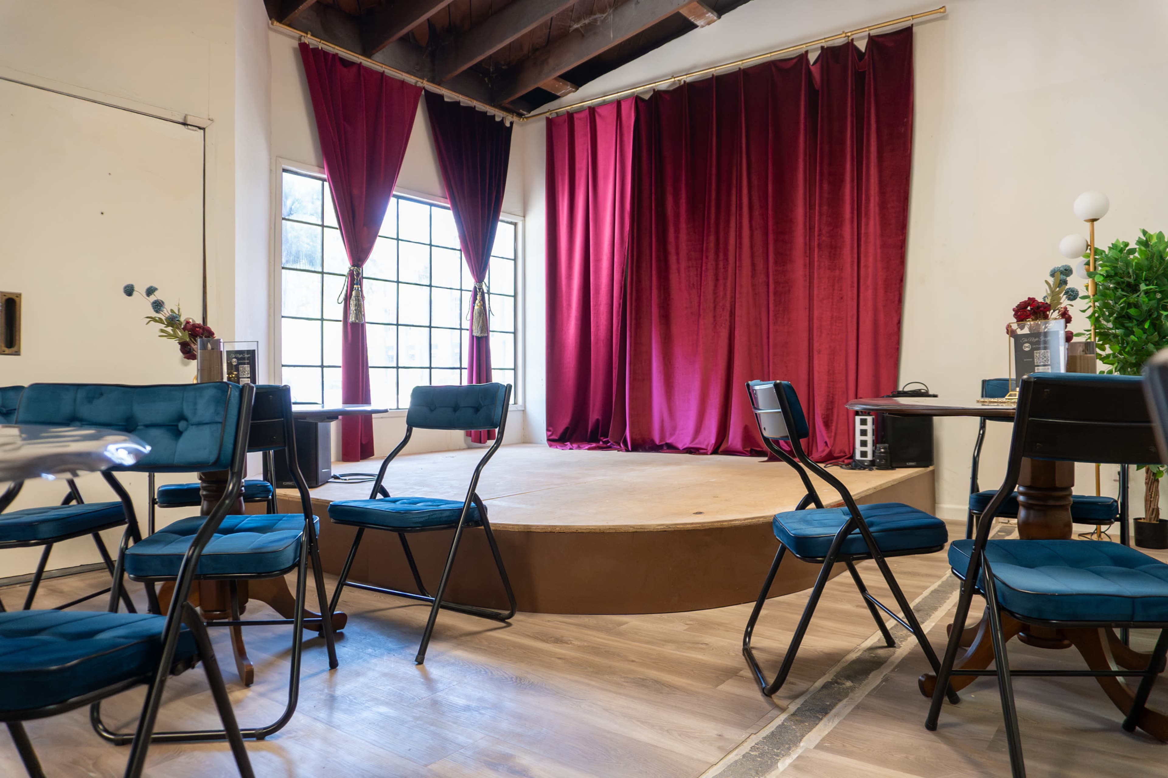 The image depicts a small stage with red curtains in a room filled with folding chairs arranged around a wooden floor platform.