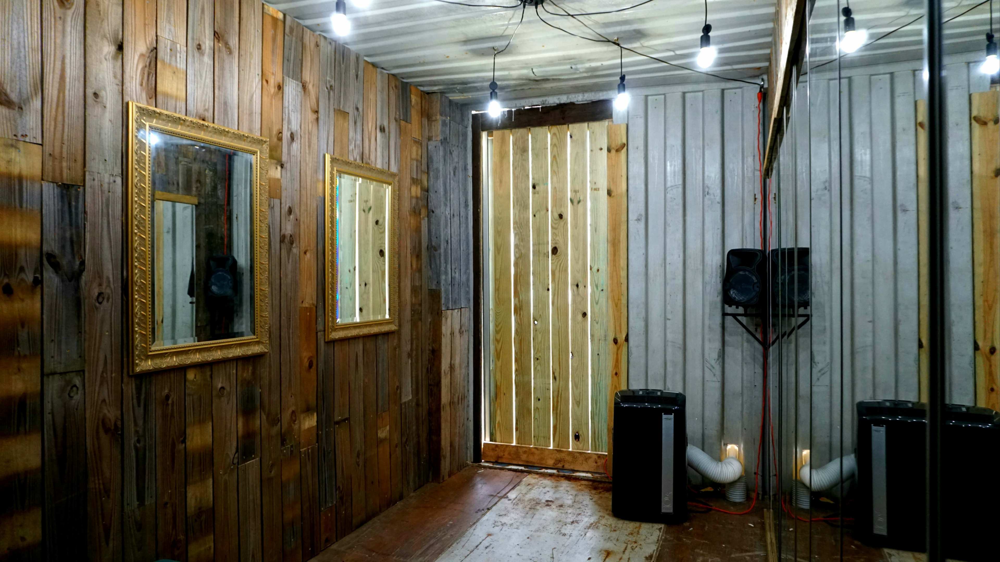 The image shows a small room with wooden walls, two framed mirrors, a wooden door, and speakers placed on the floor.