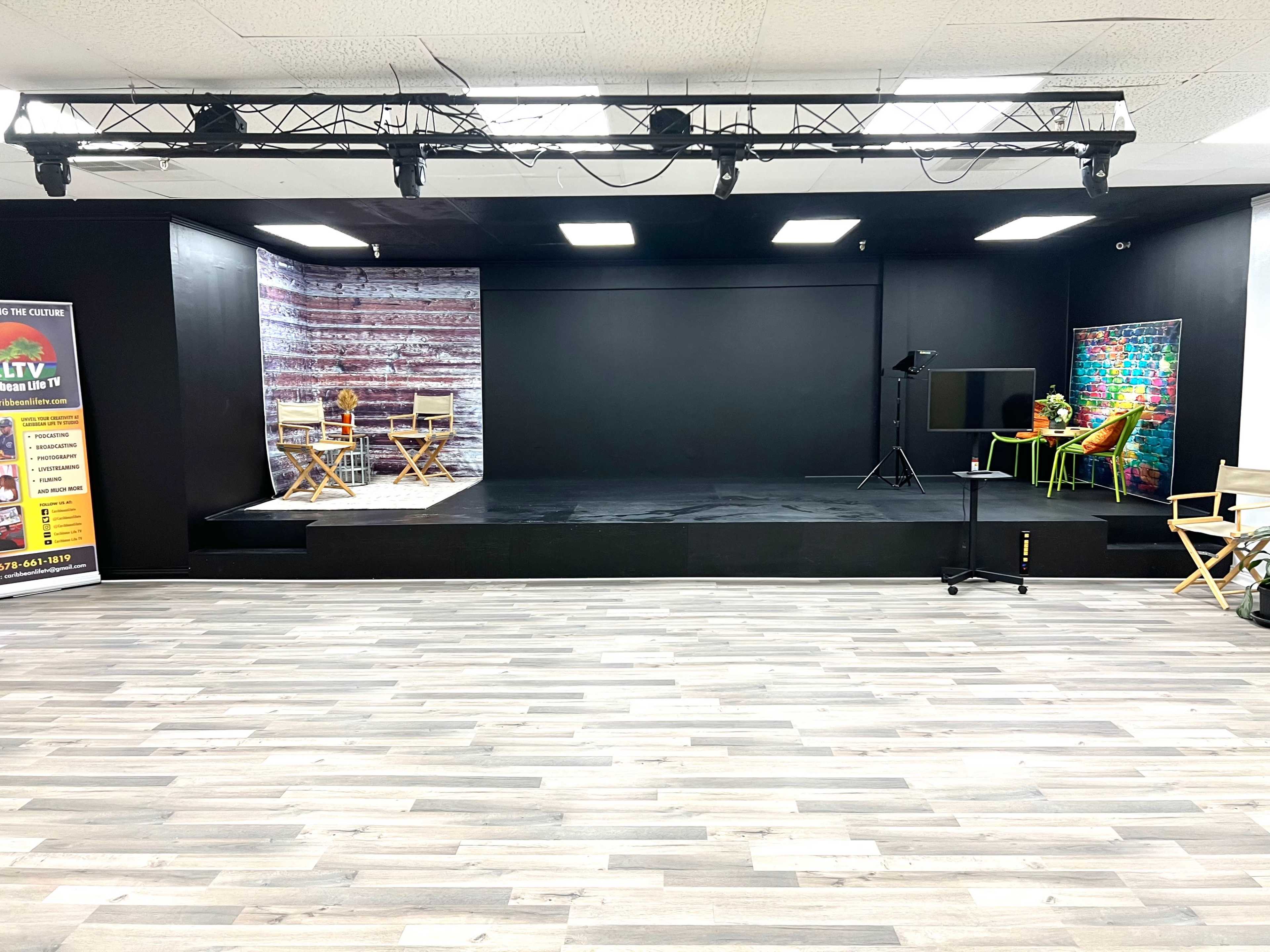 An empty studio space featuring a black back wall, wooden flooring, and areas set up with chairs and tables for potential scenes or filming.