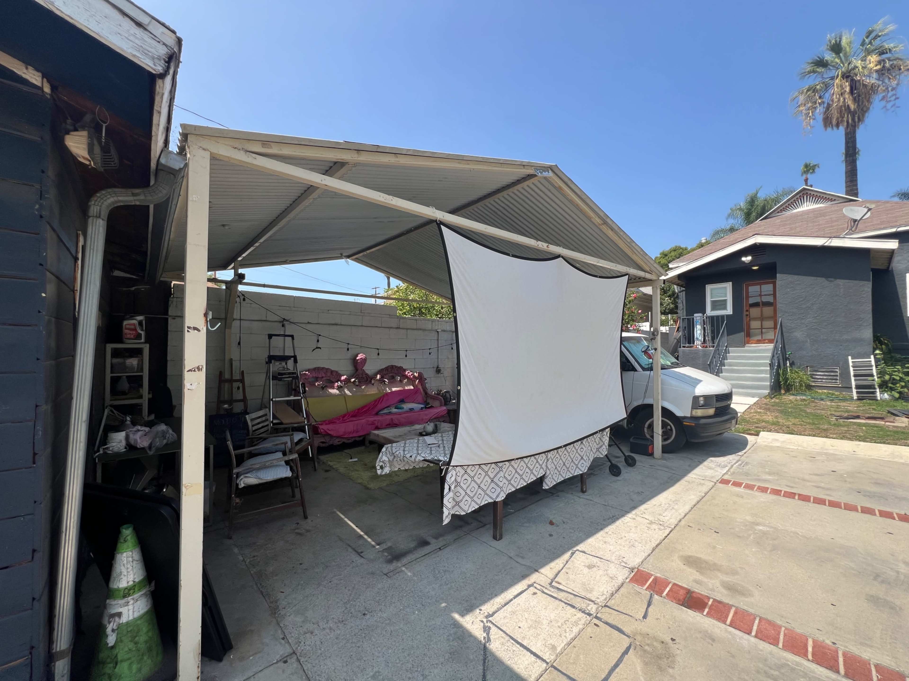 A covered outdoor area features a makeshift structure with a large white sheet, and several beds and furniture items are visible underneath it.