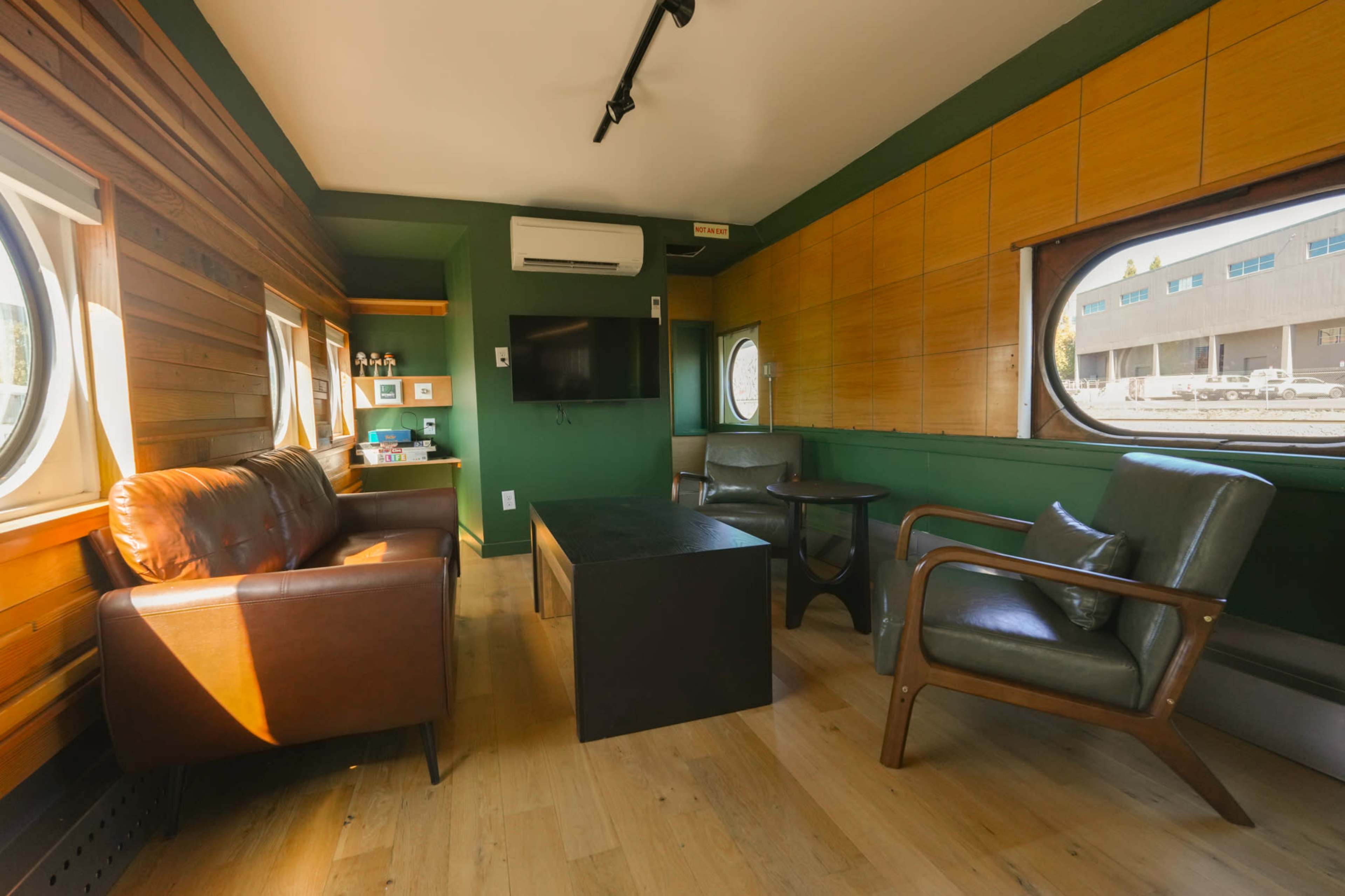 The image shows a small, modern living space with wooden walls, circular windows, a television mounted on the wall, and a seating arrangement consisting of two chairs and a leather sofa.