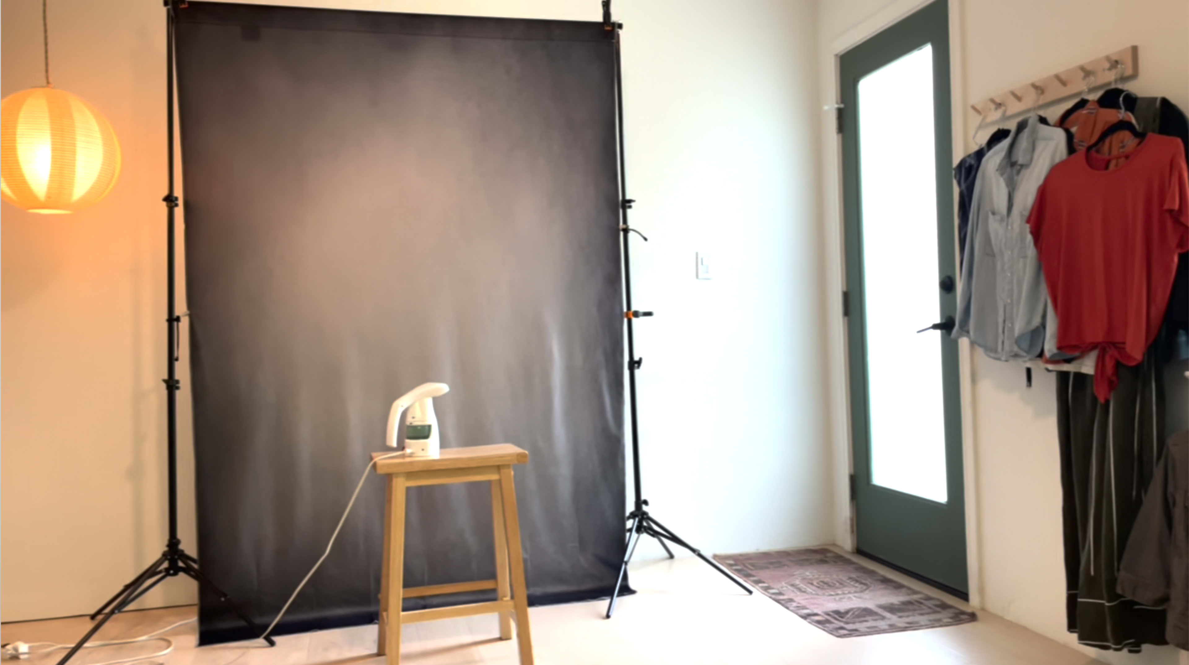 A simple indoor setup with a black backdrop, a small wooden stool, a handheld steamer, and a door with a window, alongside clothes hanging on a hook.