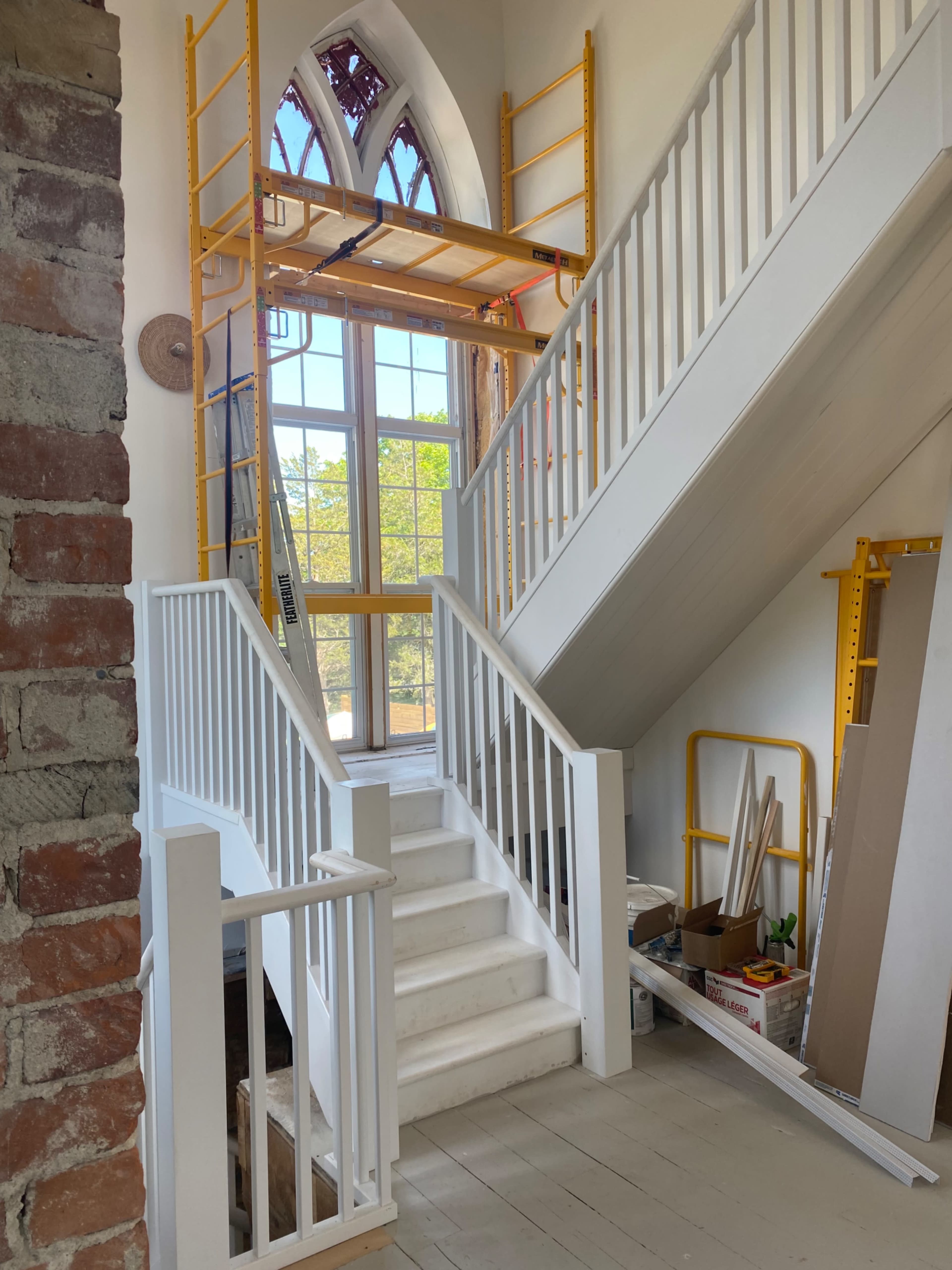 A staircase within a brightly lit interior space featuring scaffolding next to large windows and construction materials scattered around.