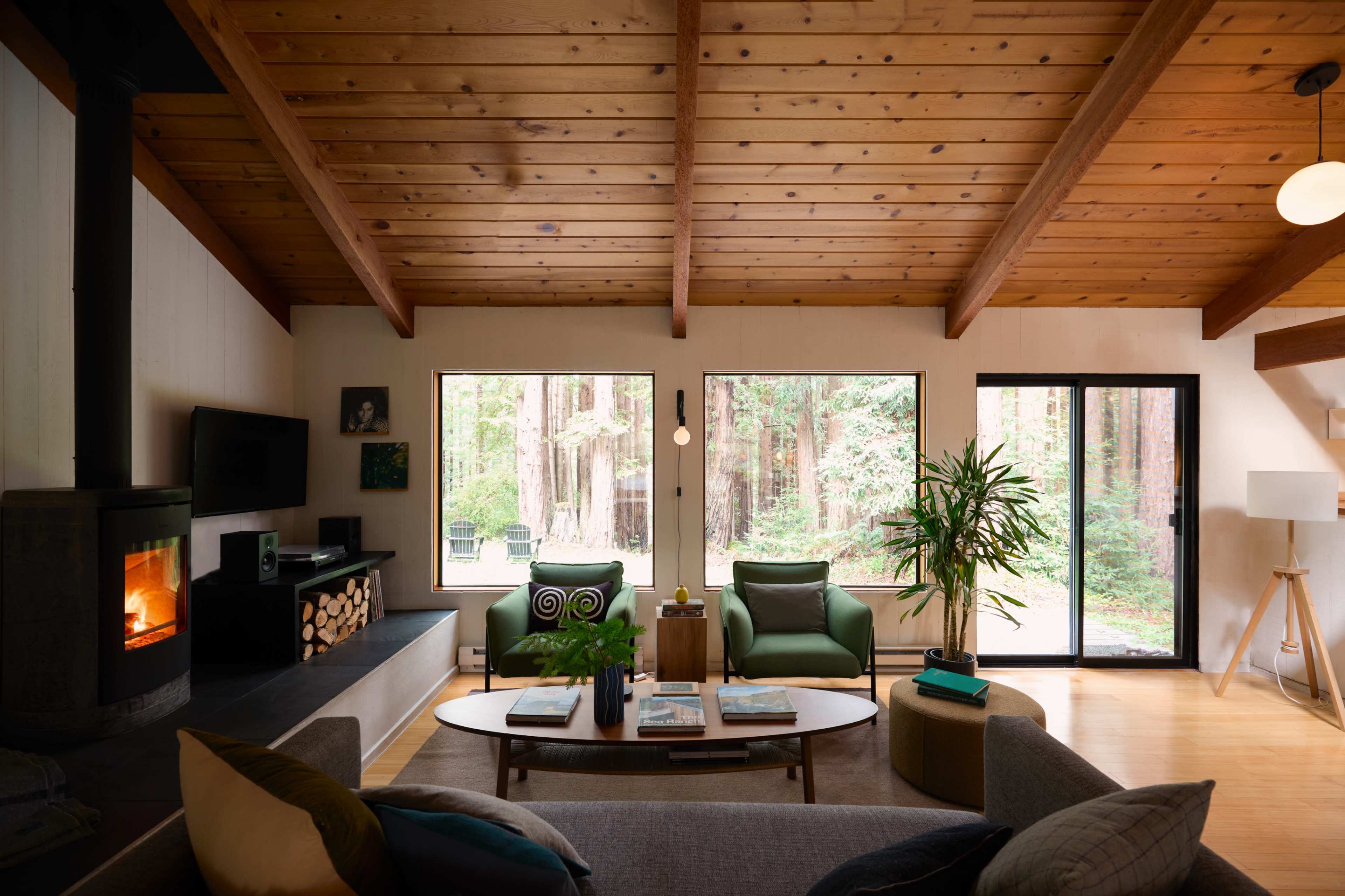The living room features large windows that overlook a forested area, showcasing a modern design with a fireplace, wooden beams, and comfortable seating.