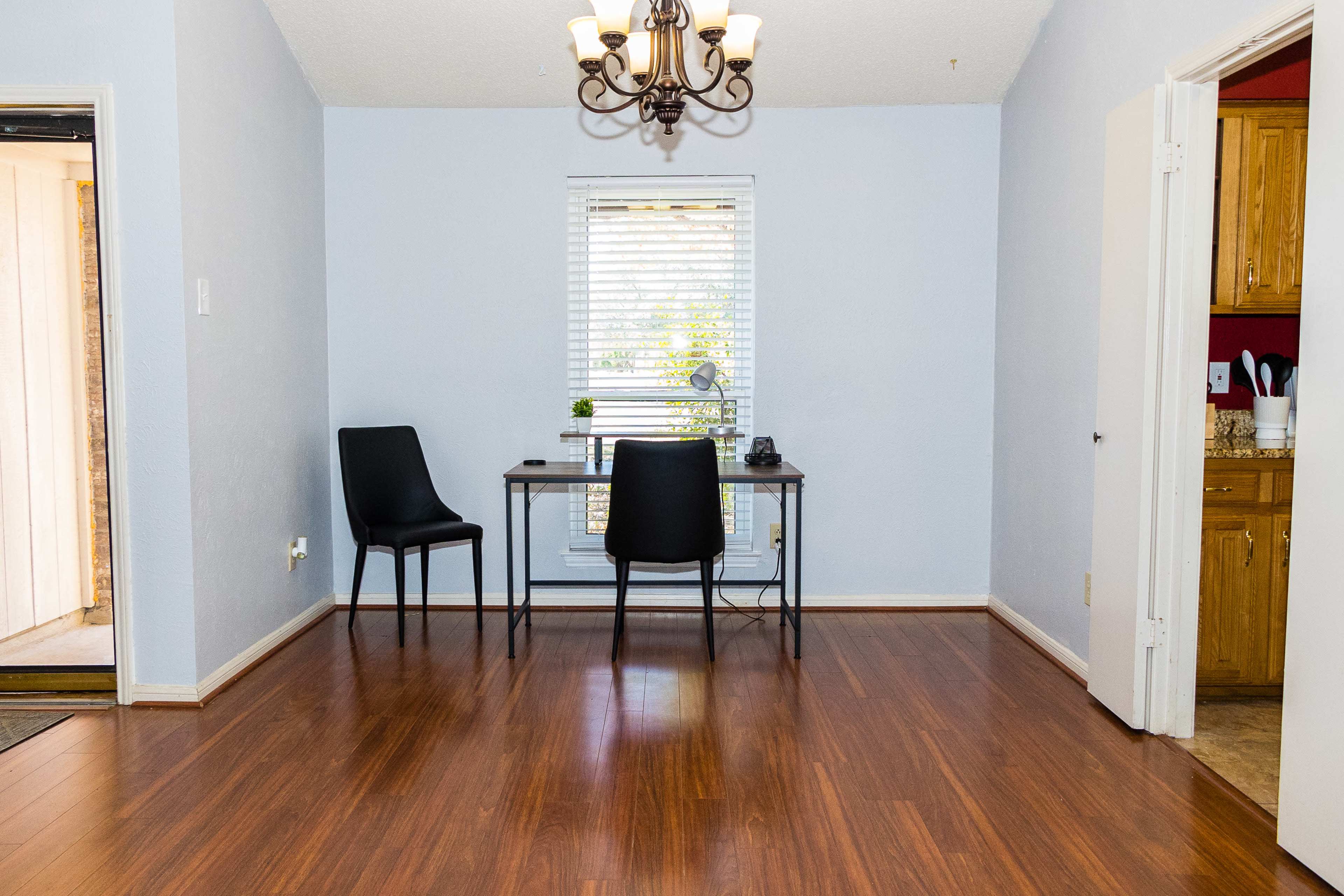 A small dining area features a window with blinds, a desk with two chairs, and wooden flooring.