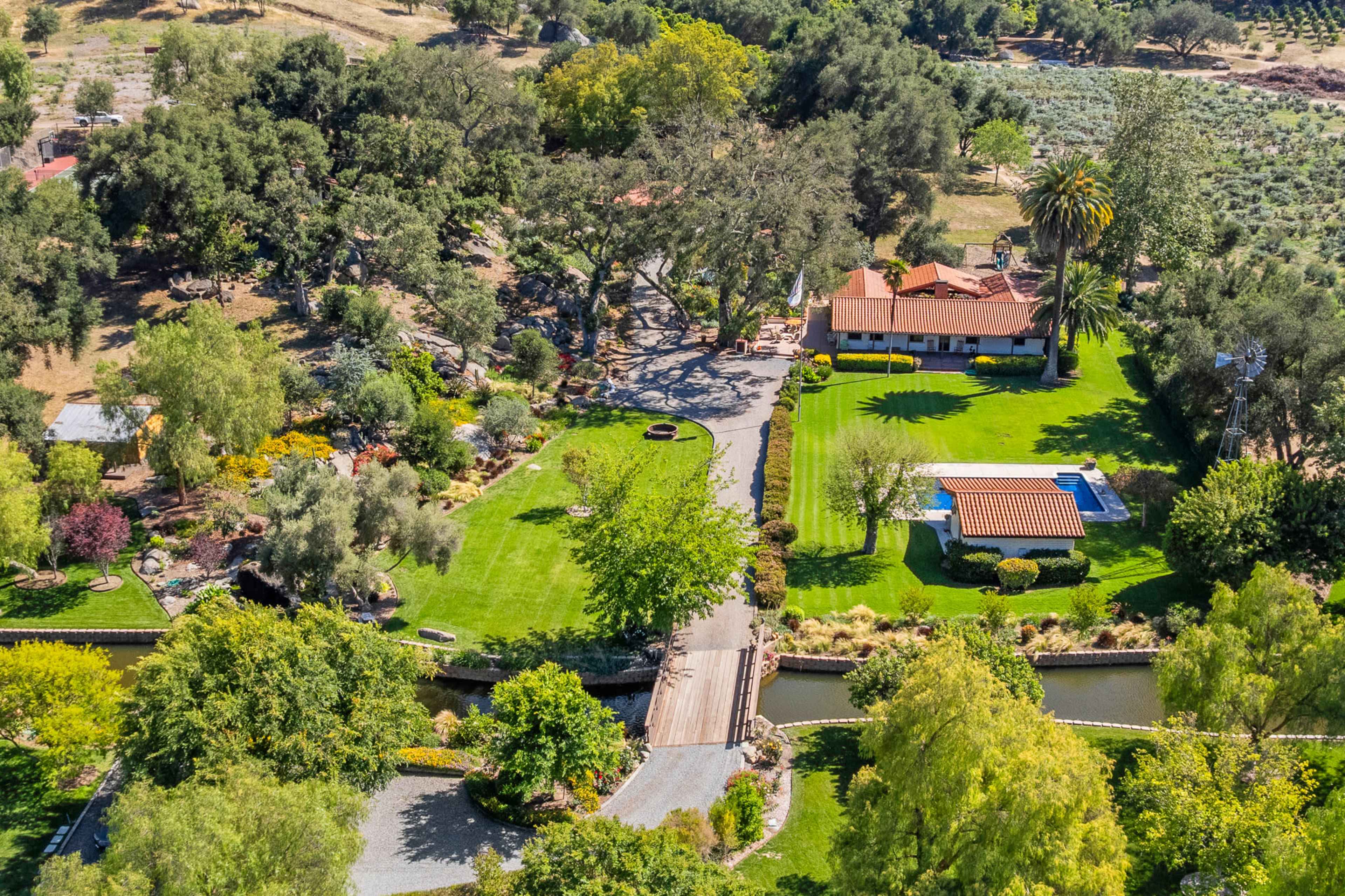 The image shows an aerial view of a landscaped property with a house, a swimming pool, gardens, and pathways surrounded by greenery and trees.