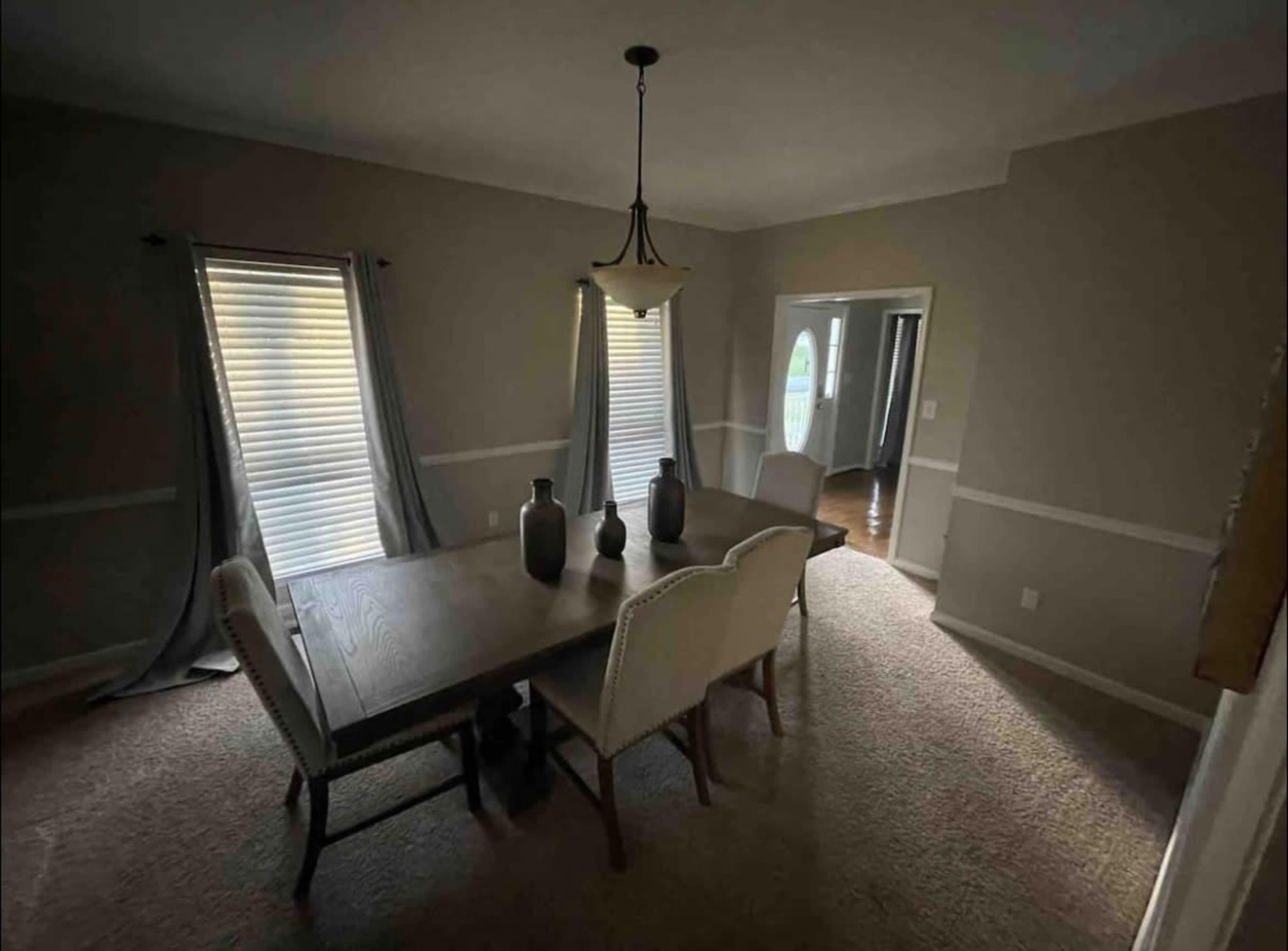 A dining room featuring a wooden table with two large vases on it, surrounded by four chairs, with two windows partially covered by curtains.