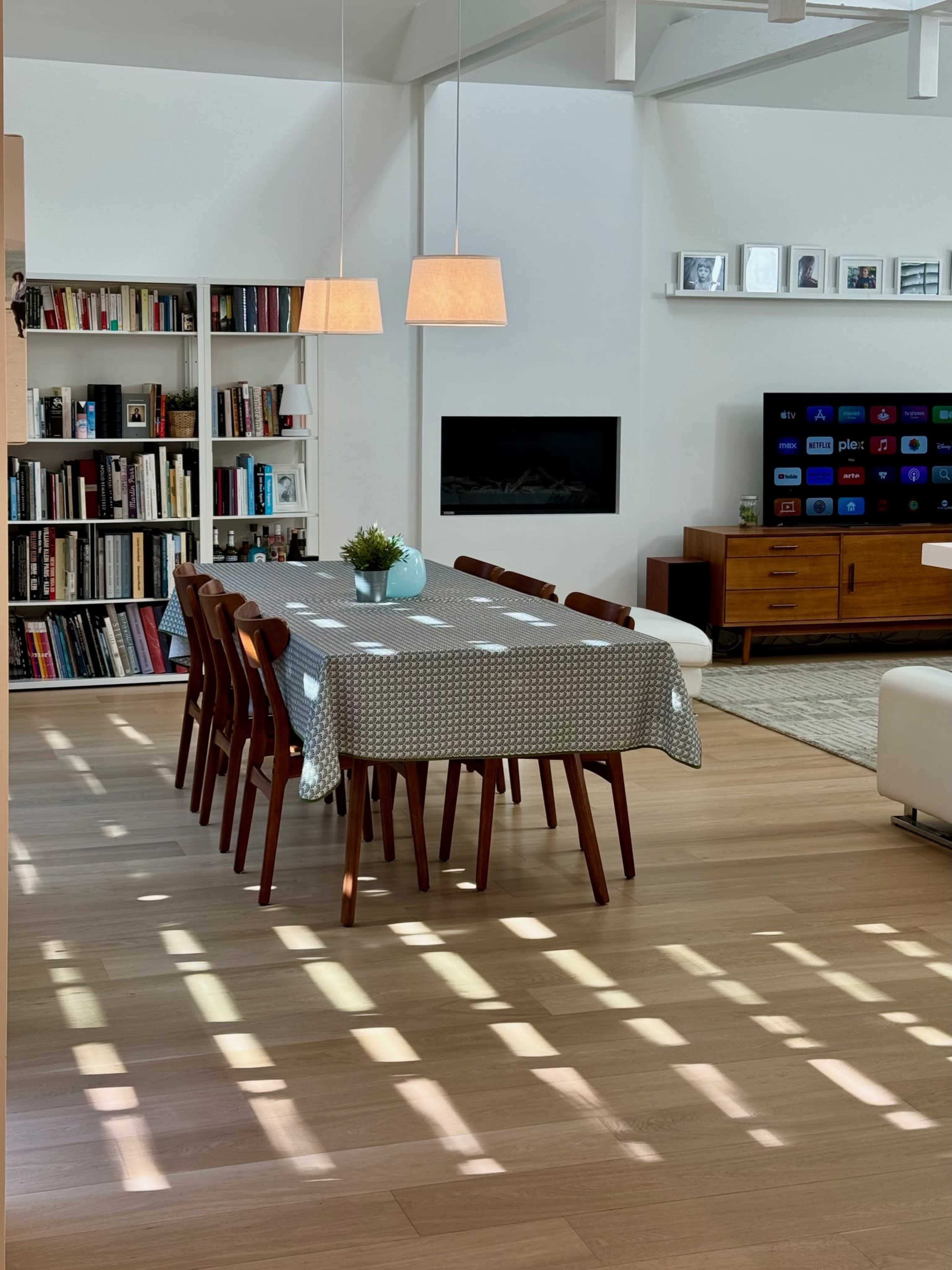 A spacious dining area features a long wooden table with a light-gray tablecloth, surrounded by eight chairs, in front of a wall-mounted television and a bookshelf.