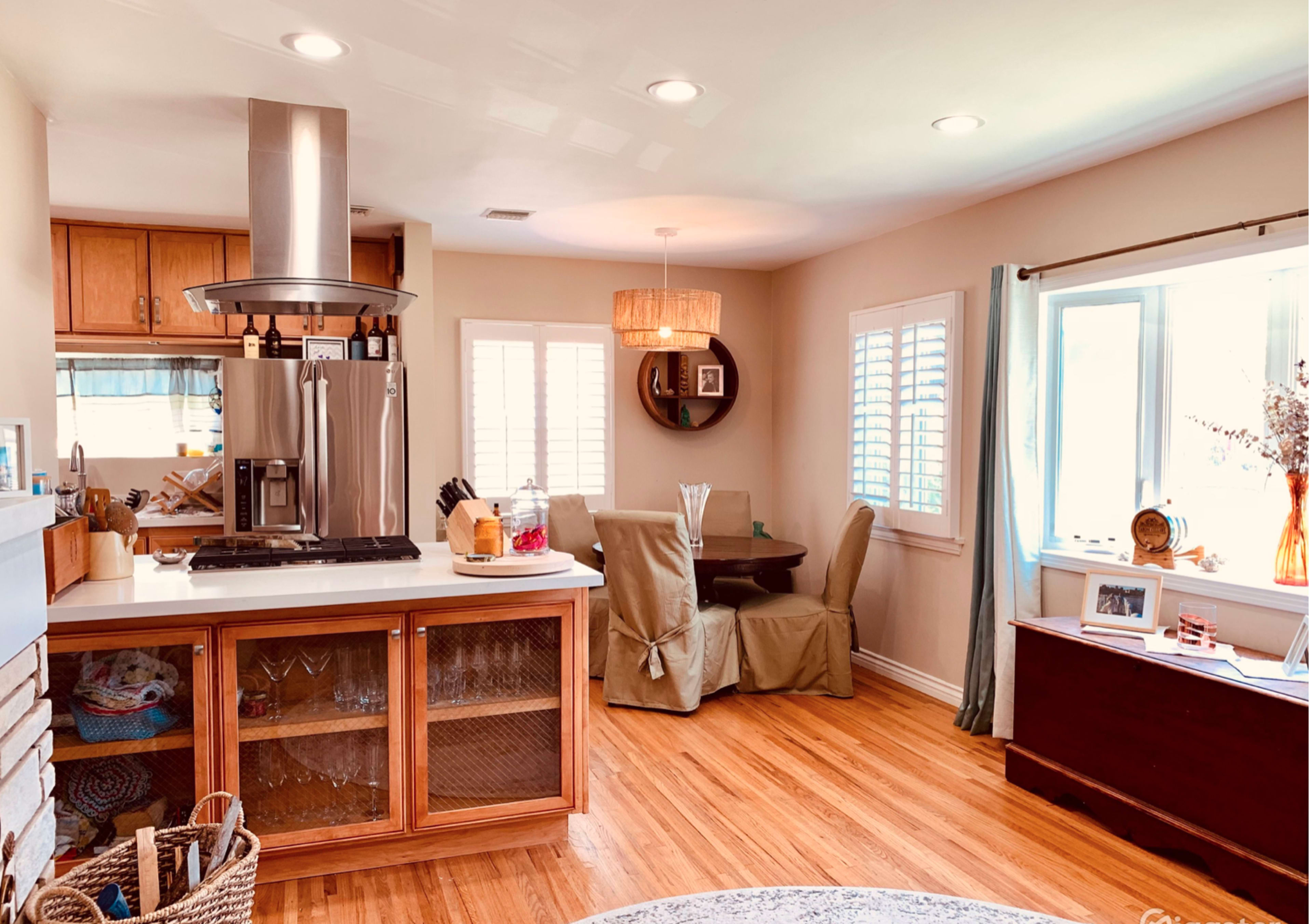 The image shows a bright kitchen and dining area with wooden cabinetry, a stainless steel refrigerator, and a round table set for dining.