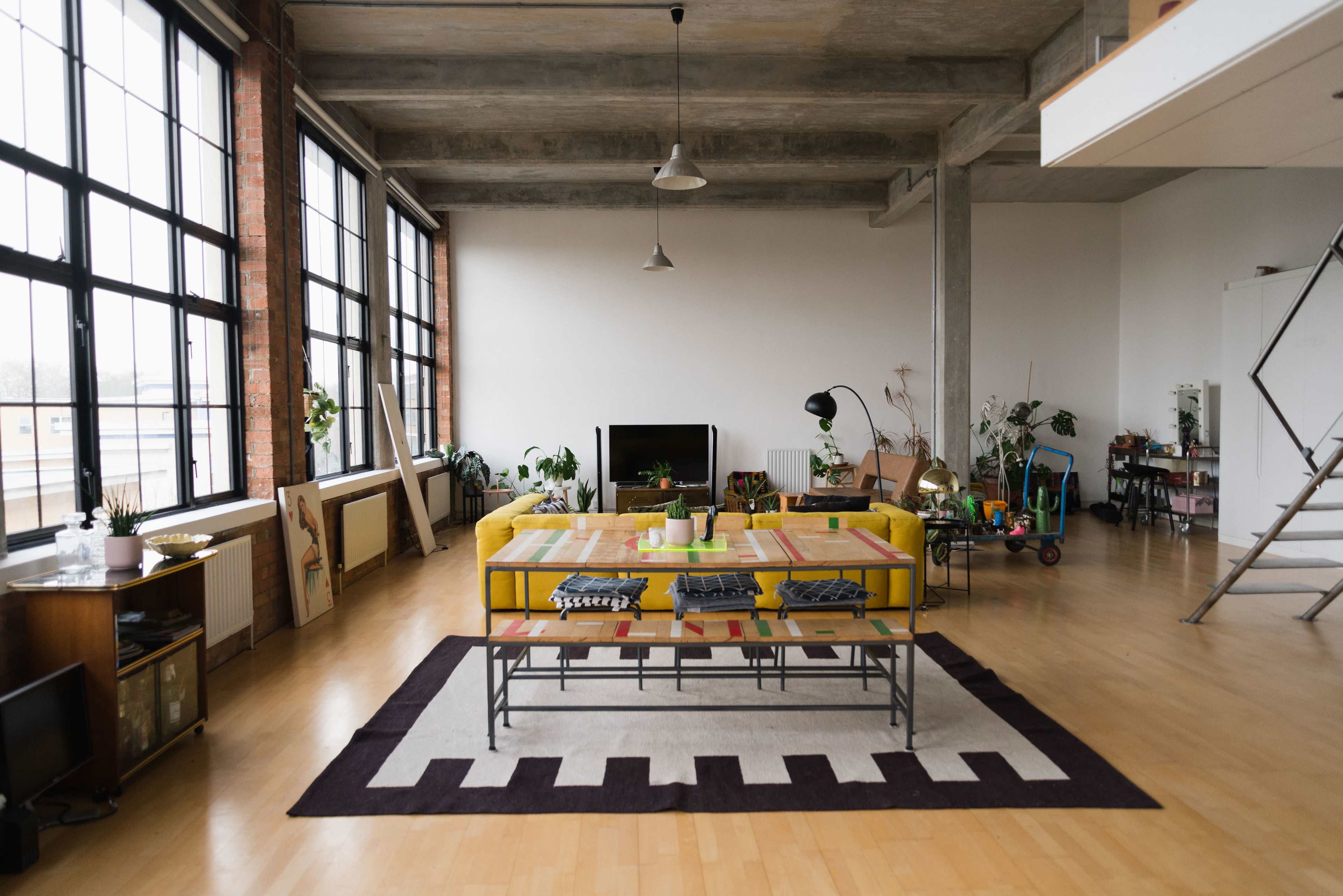 A spacious living area features large windows, a colorful couch, a coffee table, and various indoor plants.
