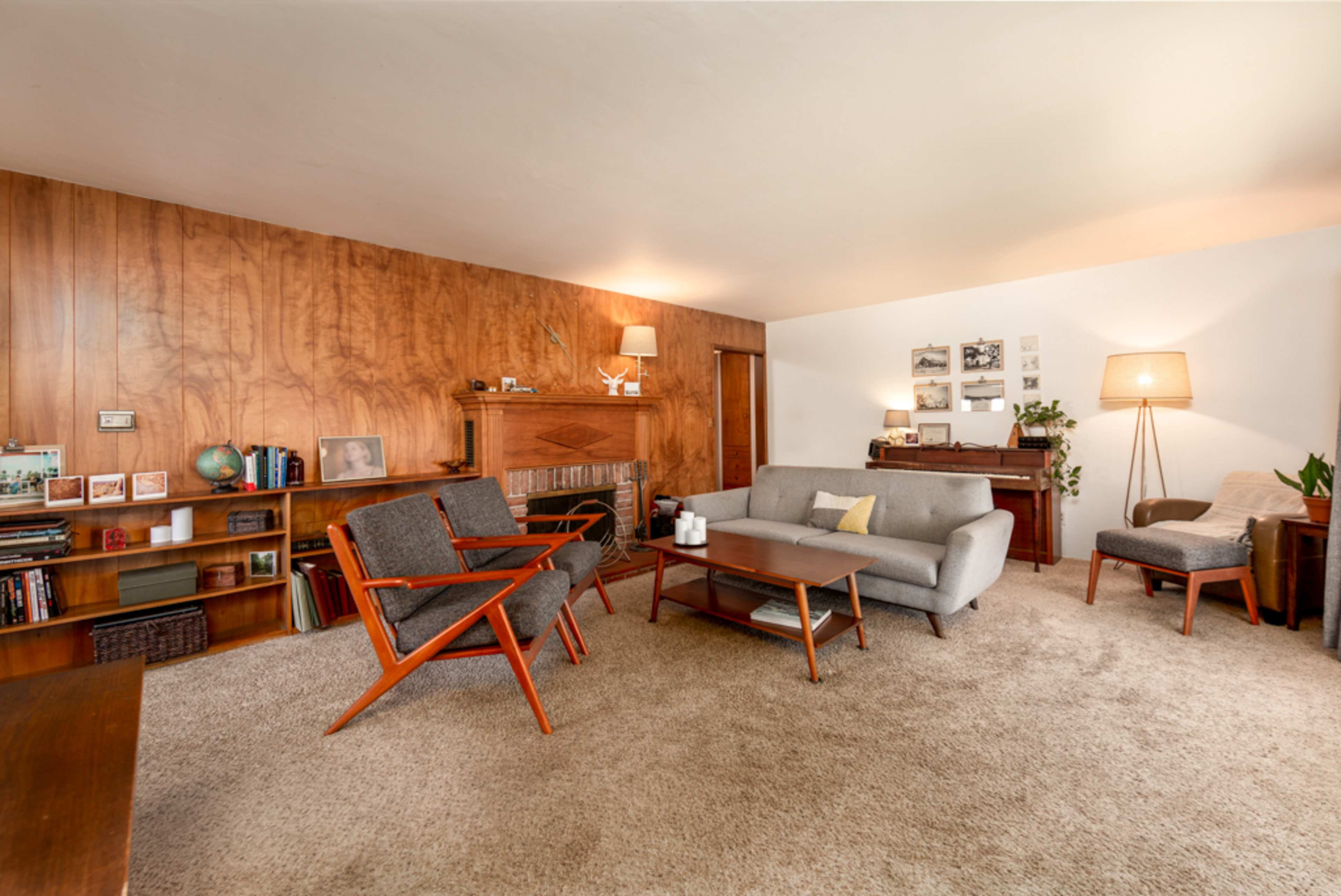 A mid-century modern living room with wooden paneling, a gray sofa, vintage chairs, and a styled bookshelf against one wall.