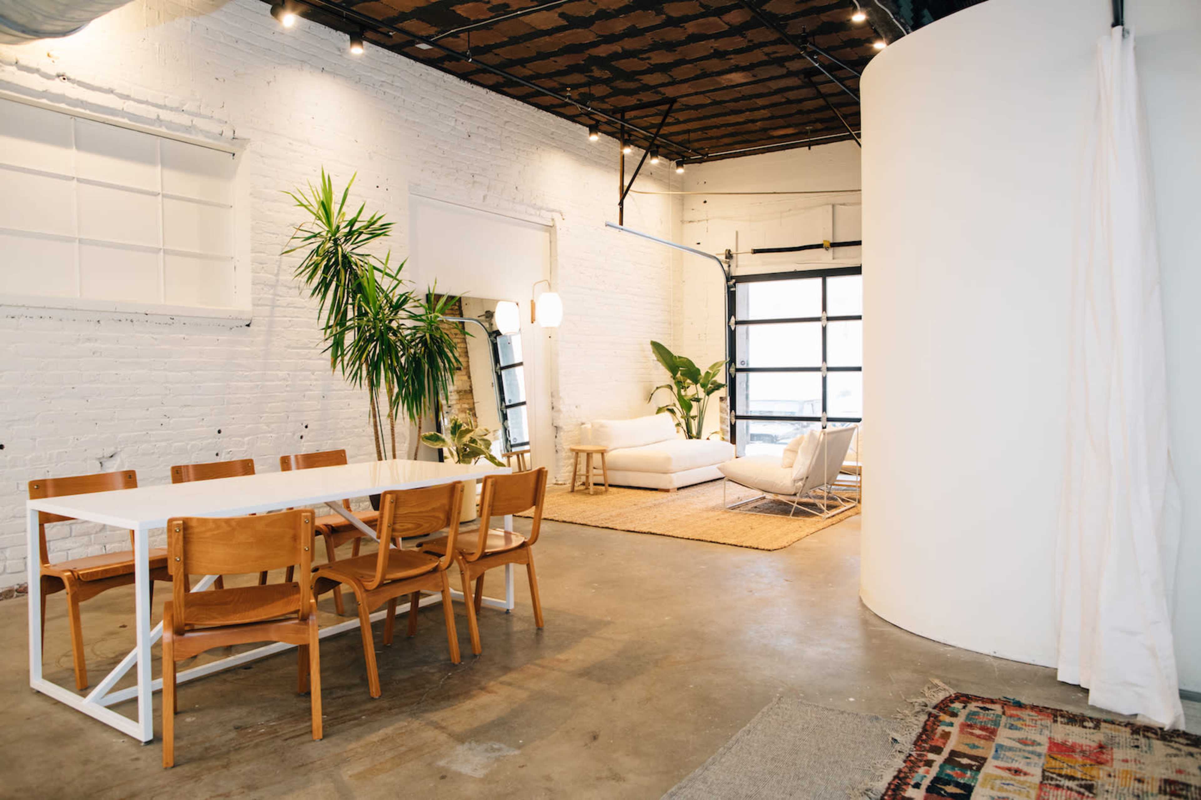 The image shows a spacious, modern interior featuring a dining area with a table and chairs, a living space with a sofa, and a large window.