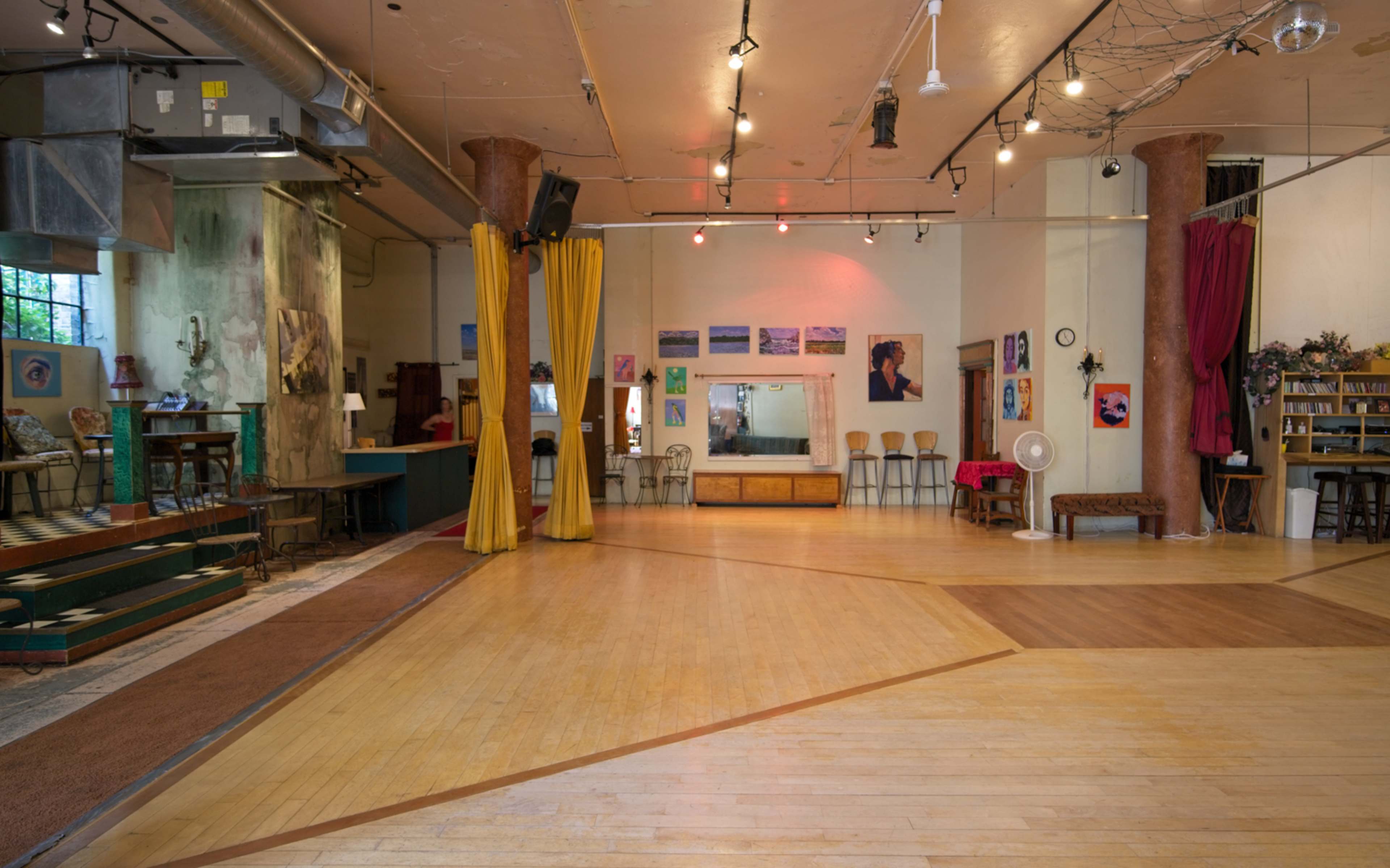 An empty dance studio with wooden flooring, colorful curtains, and framed artworks on the walls.
