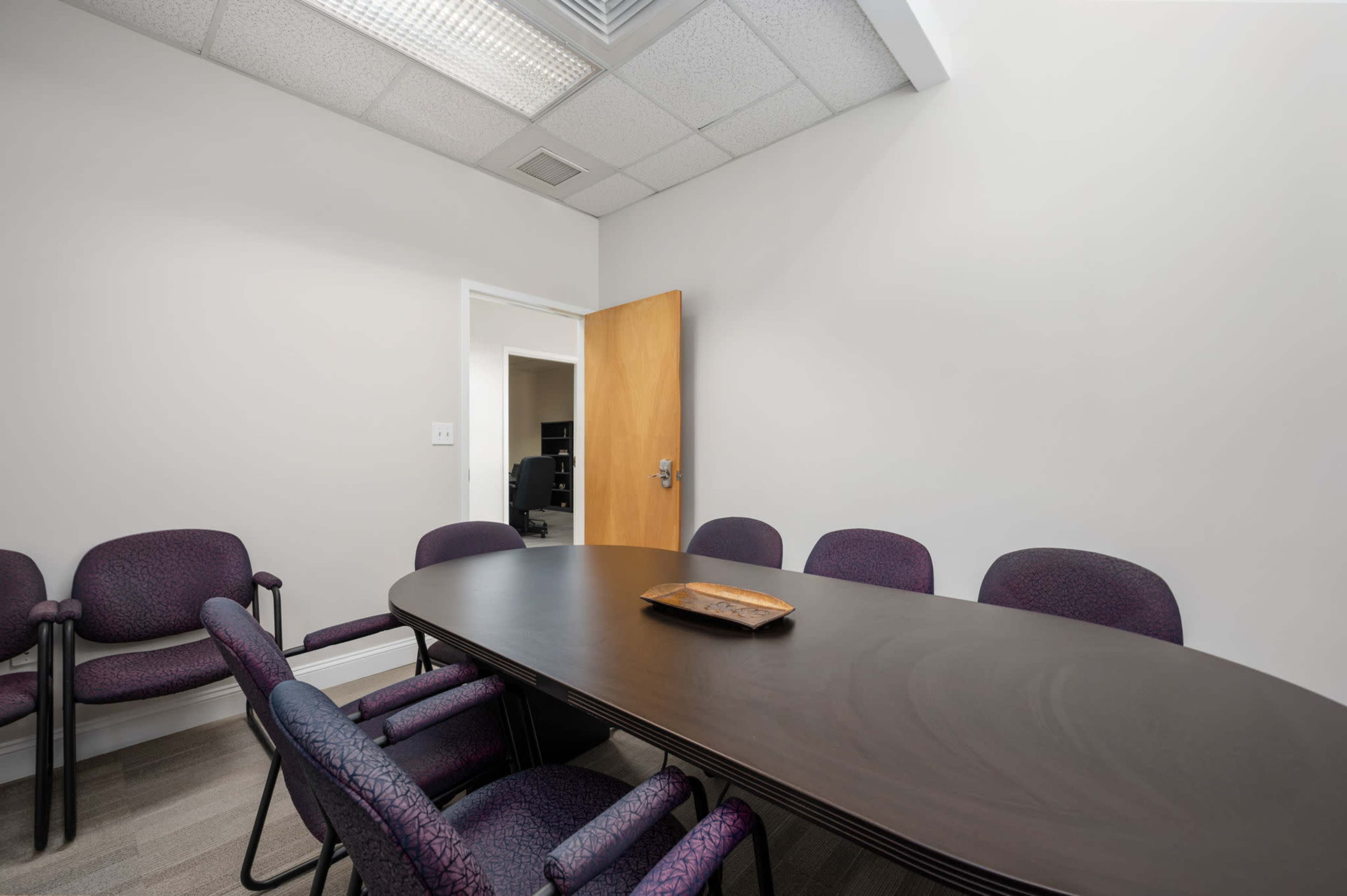 The image shows a small conference room with a circular table and several purple chairs arranged around it, with a wooden door and a glimpse of an office beyond.