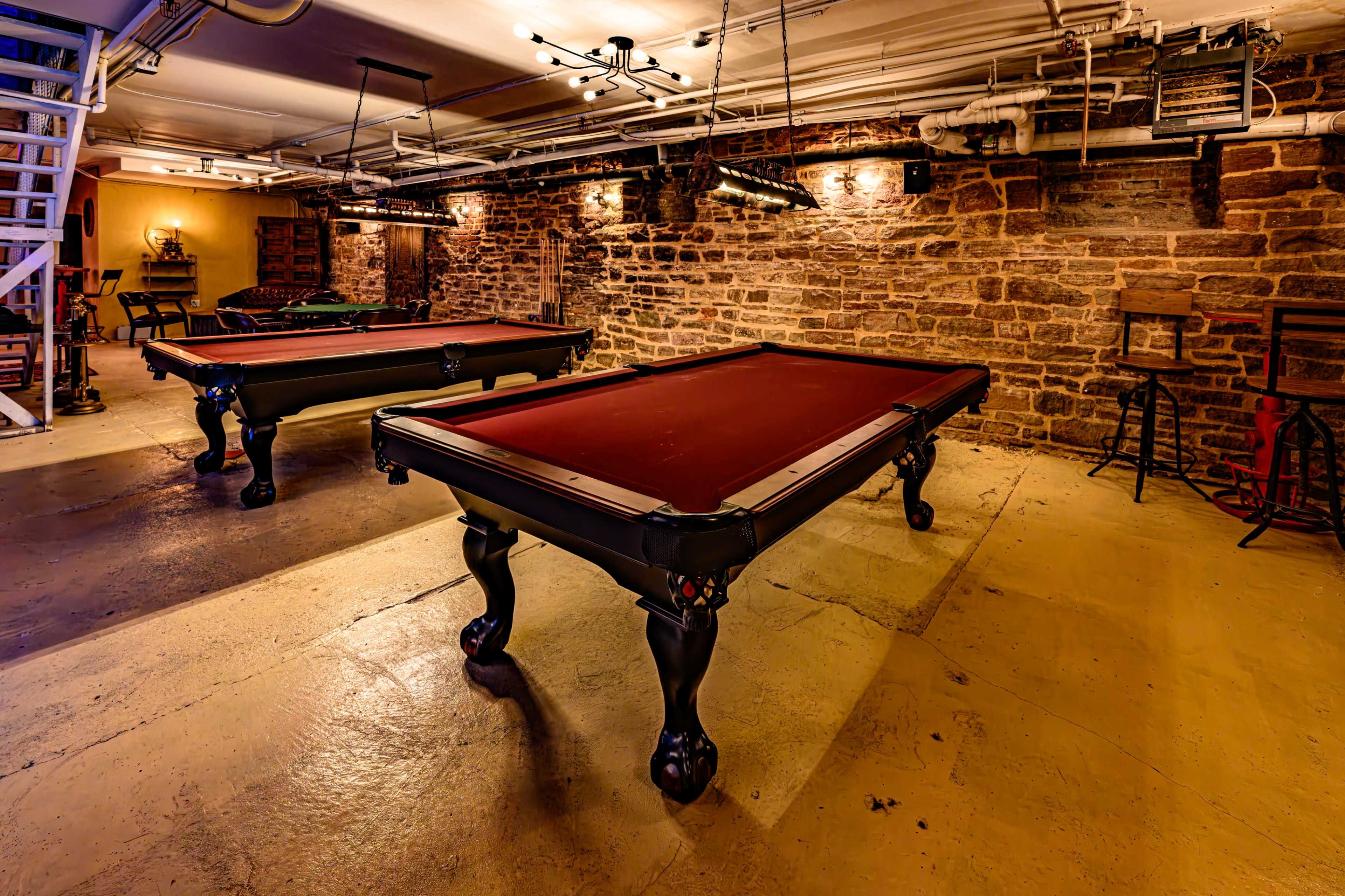 The image shows a game room featuring two pool tables on a concrete floor with exposed stone walls and overhead lighting.