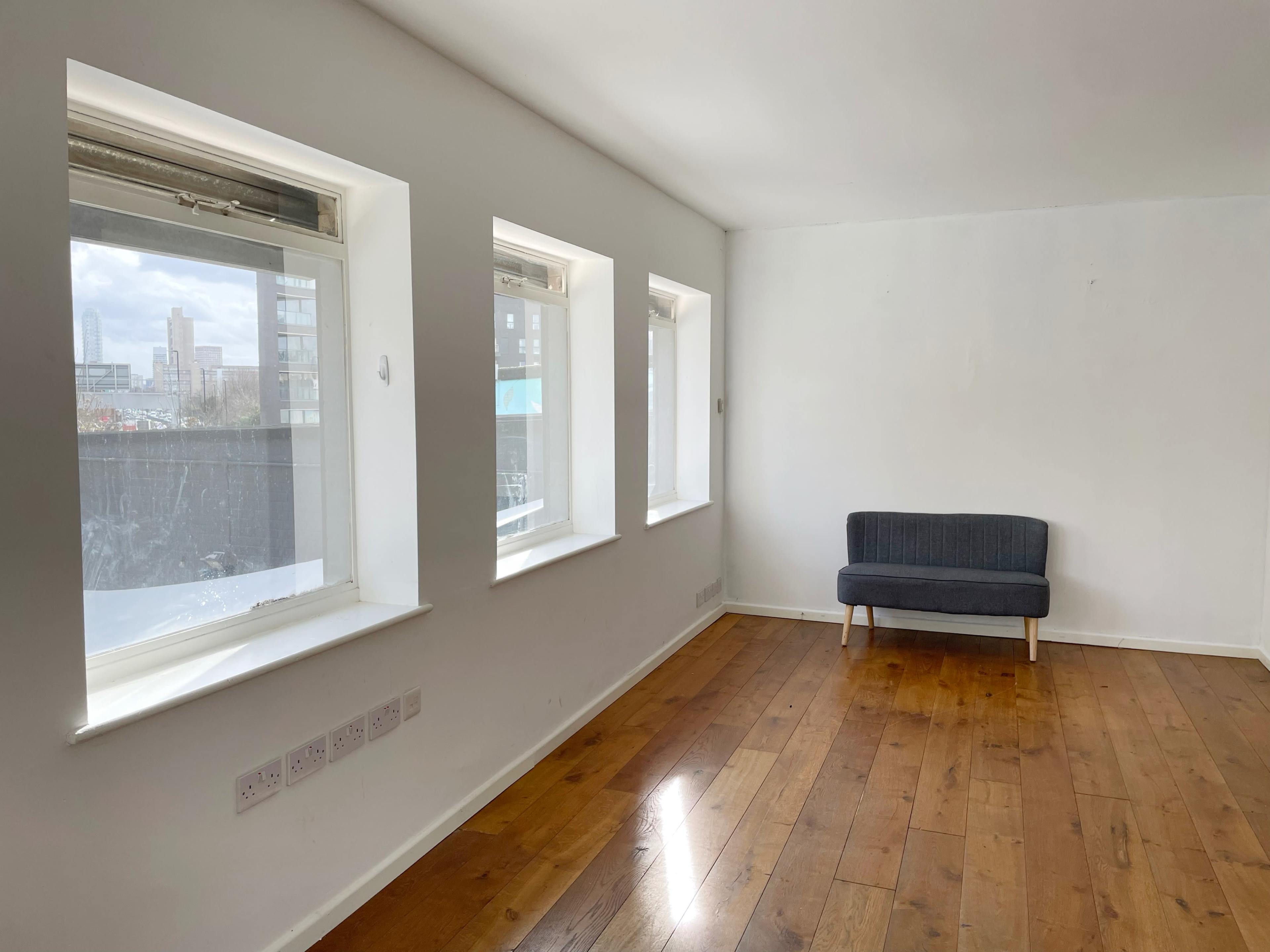 A room with three windows and a gray couch against a white wall, featuring hardwood flooring.