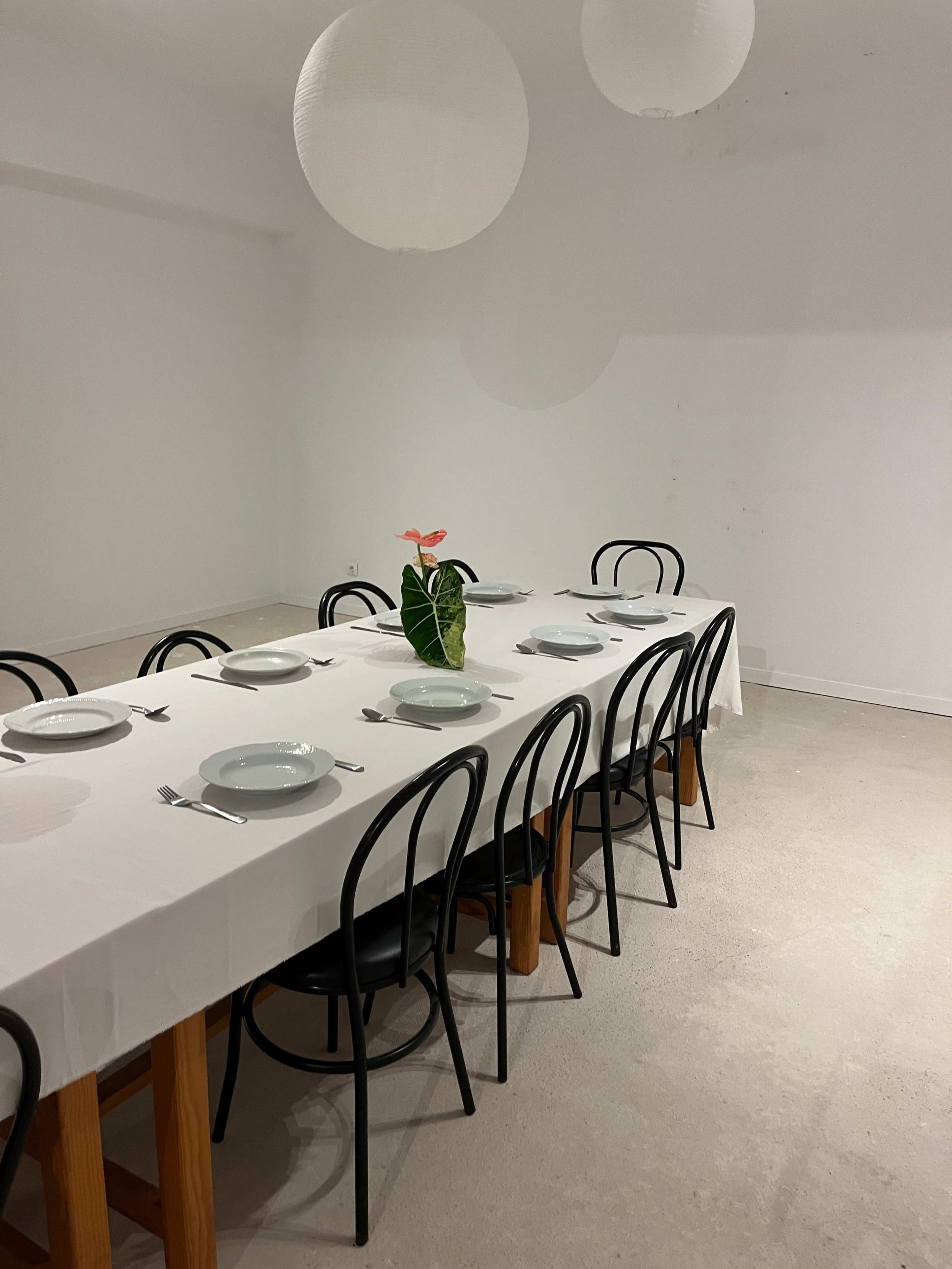 A long rectangular table is set with plates and cutlery, surrounded by black chairs, with a plant centerpiece and paper lanterns hanging above.