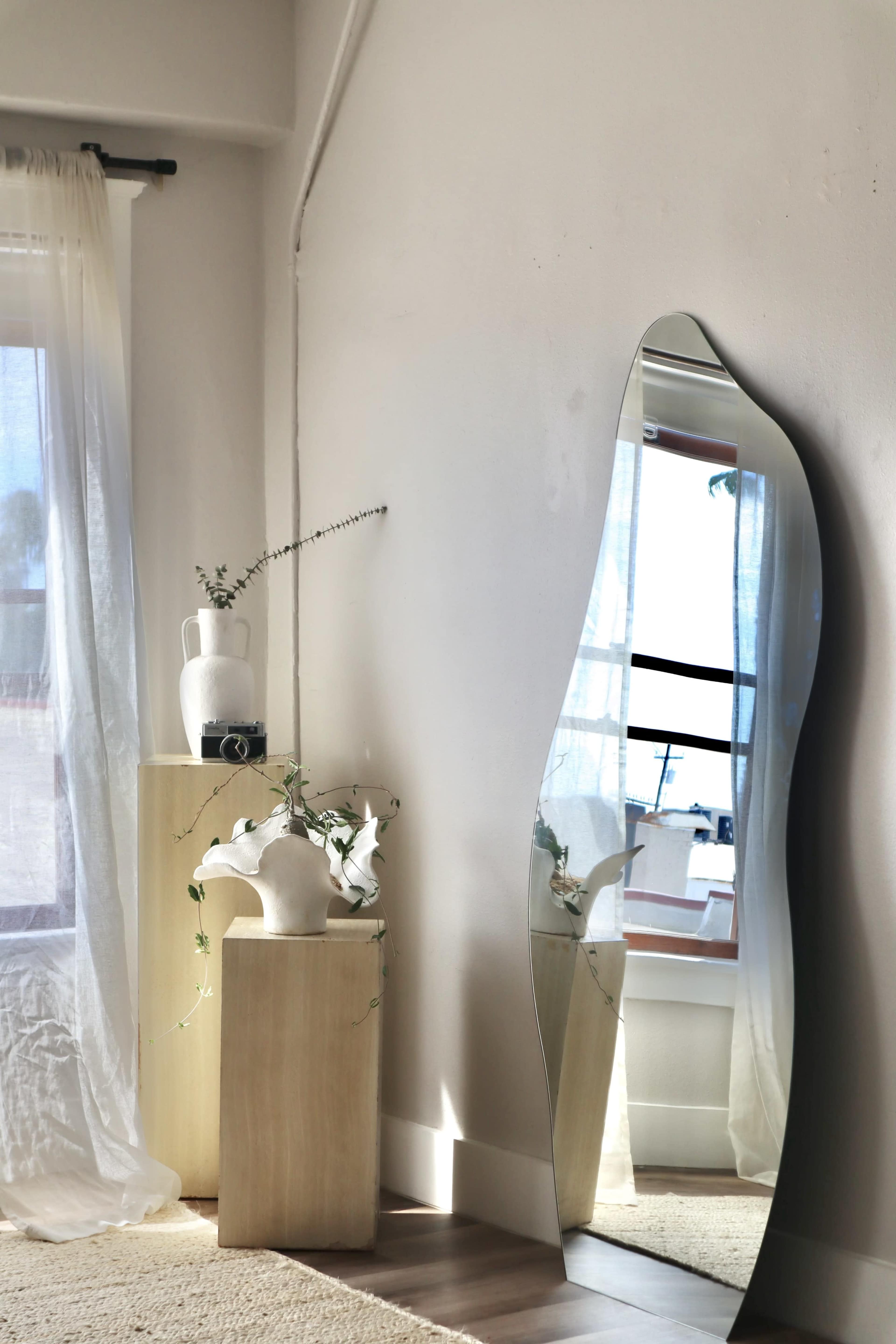 A tall, wavy mirror leans against a wall beside two light-colored pedestals, one holding a ceramic vase and the other a white pitcher with flowers.