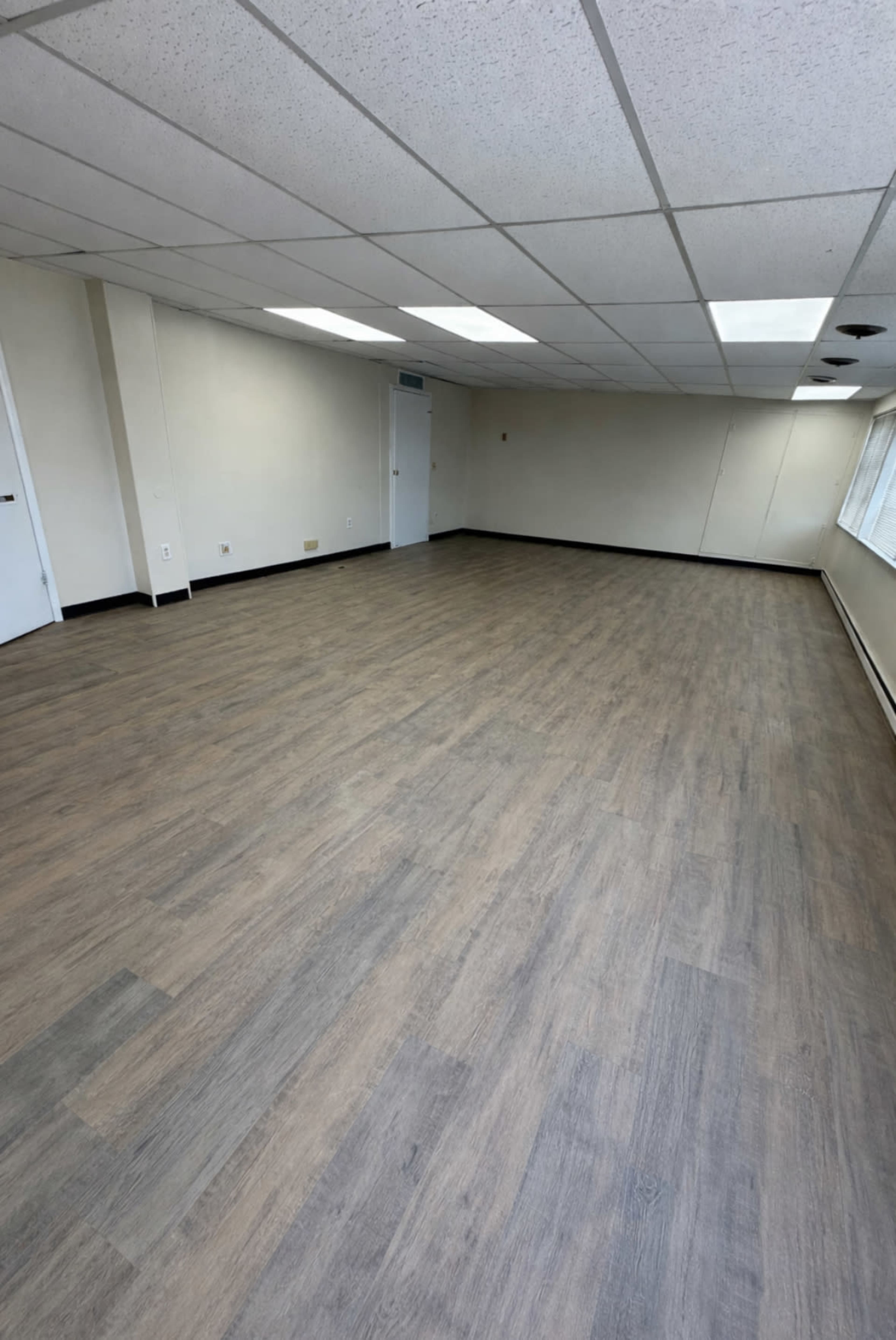 The image shows an empty room with a light-colored wooden floor, white walls, and a ceiling with fluorescent lights.