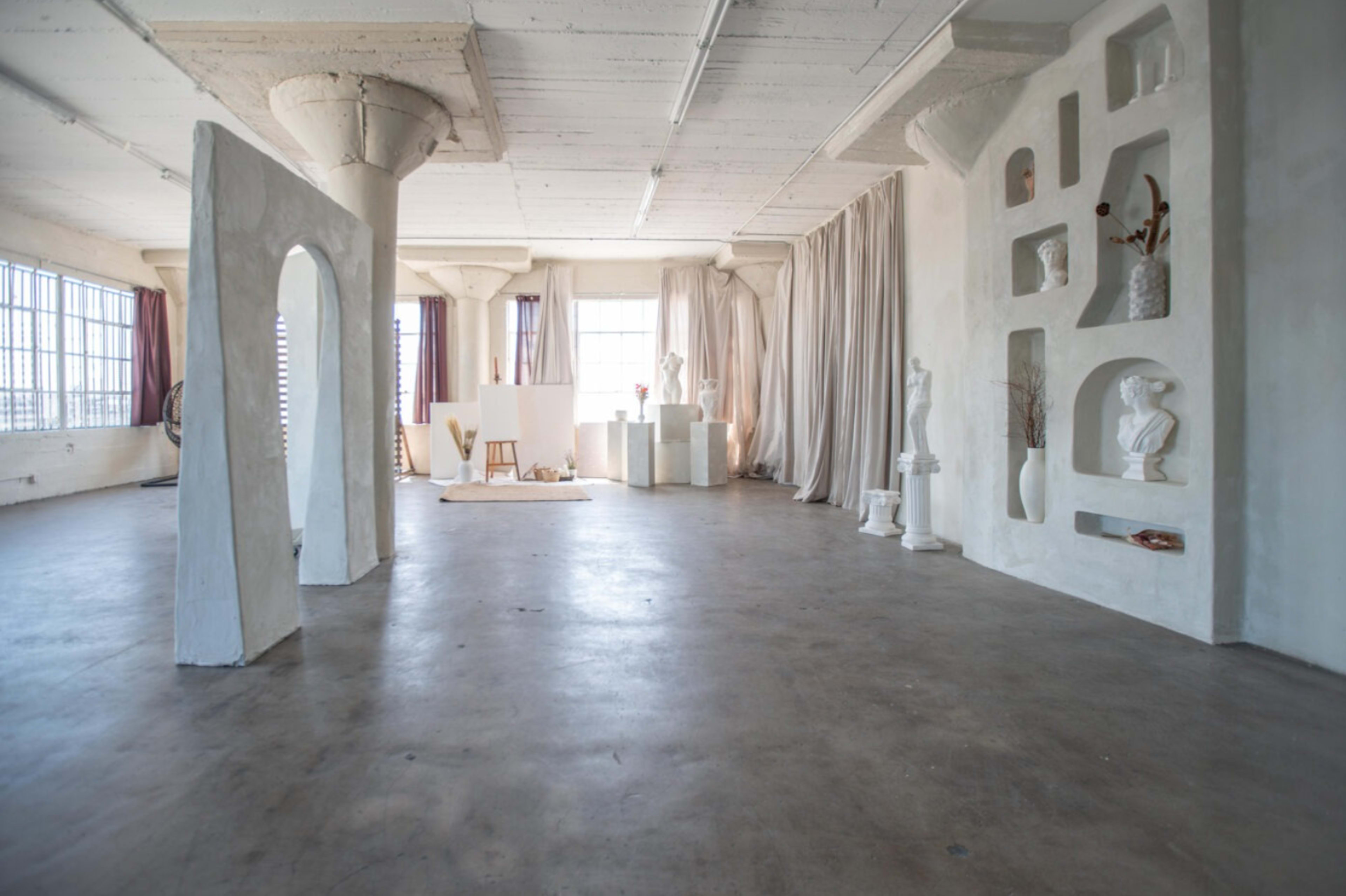 A spacious, minimally decorated interior with white walls, concrete flooring, and various sculptures arranged throughout.