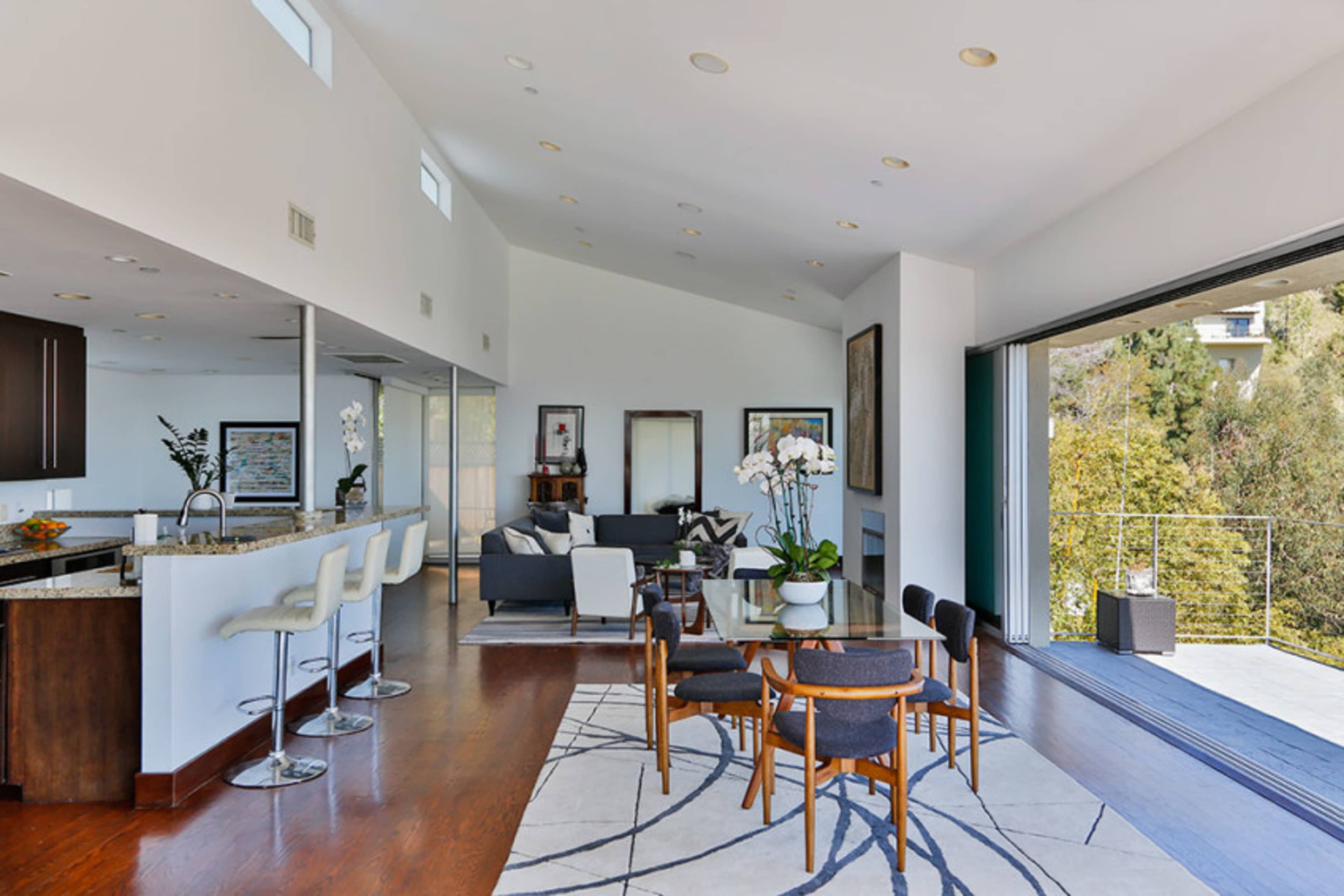A modern open-plan living space featuring a dining area with a large table, surrounded by chairs, a kitchen with bar seating, and large sliding doors opening to an outdoor area.