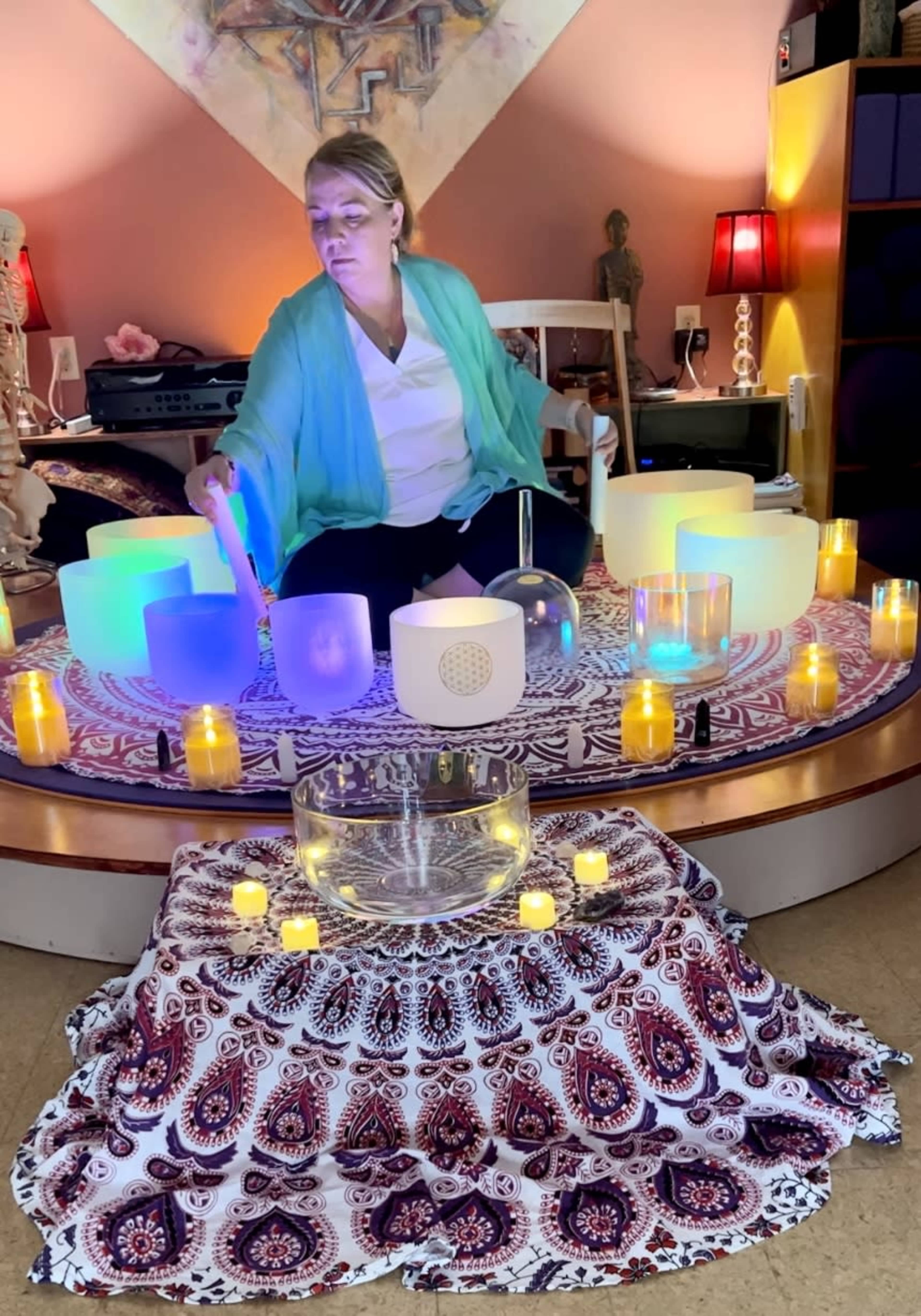 A woman in a light blue shawl sits on a patterned rug, surrounded by crystal singing bowls and lit candles, while preparing for a sound healing session.