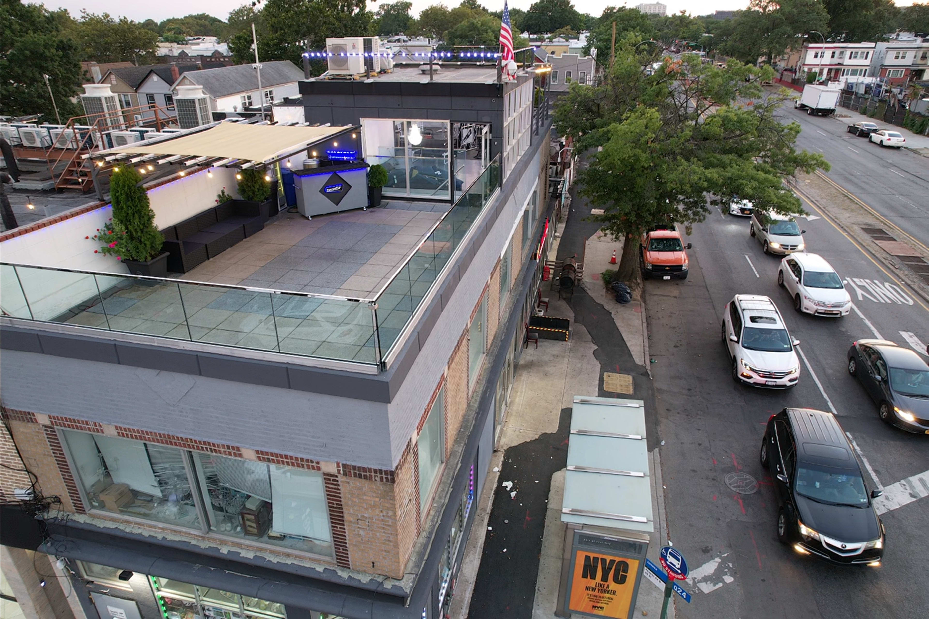 The image shows a multi-level building with a rooftop area and a view of a busy street lined with parked cars and trees.