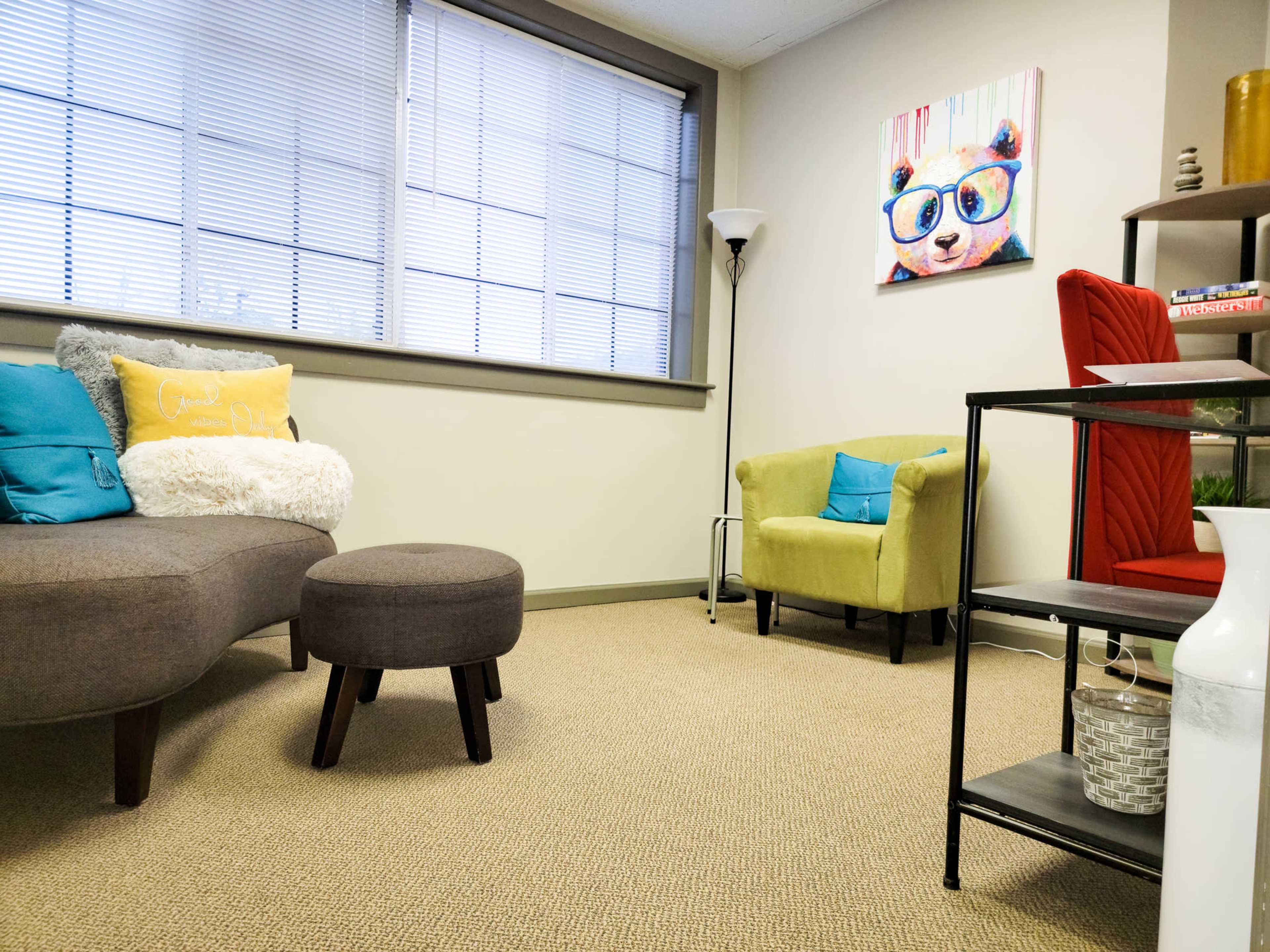 A cozy room with two armchairs, a couch, and a small round ottoman, all arranged on a carpeted floor beneath bright windows.