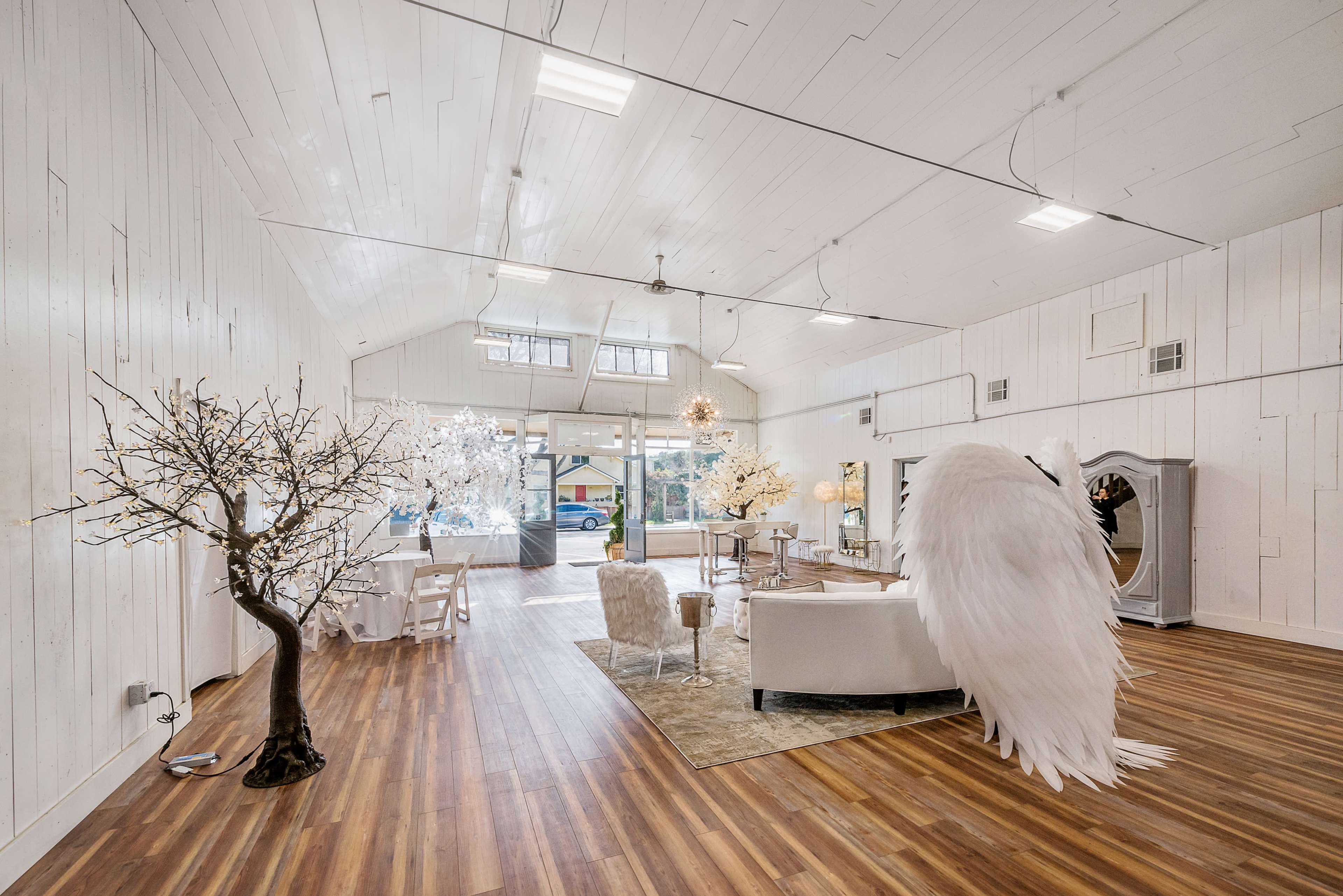 The interior of a spacious event venue features white walls, wooden flooring, a large arrangement of white decorations, and a prominent installation resembling a bird with large wings.