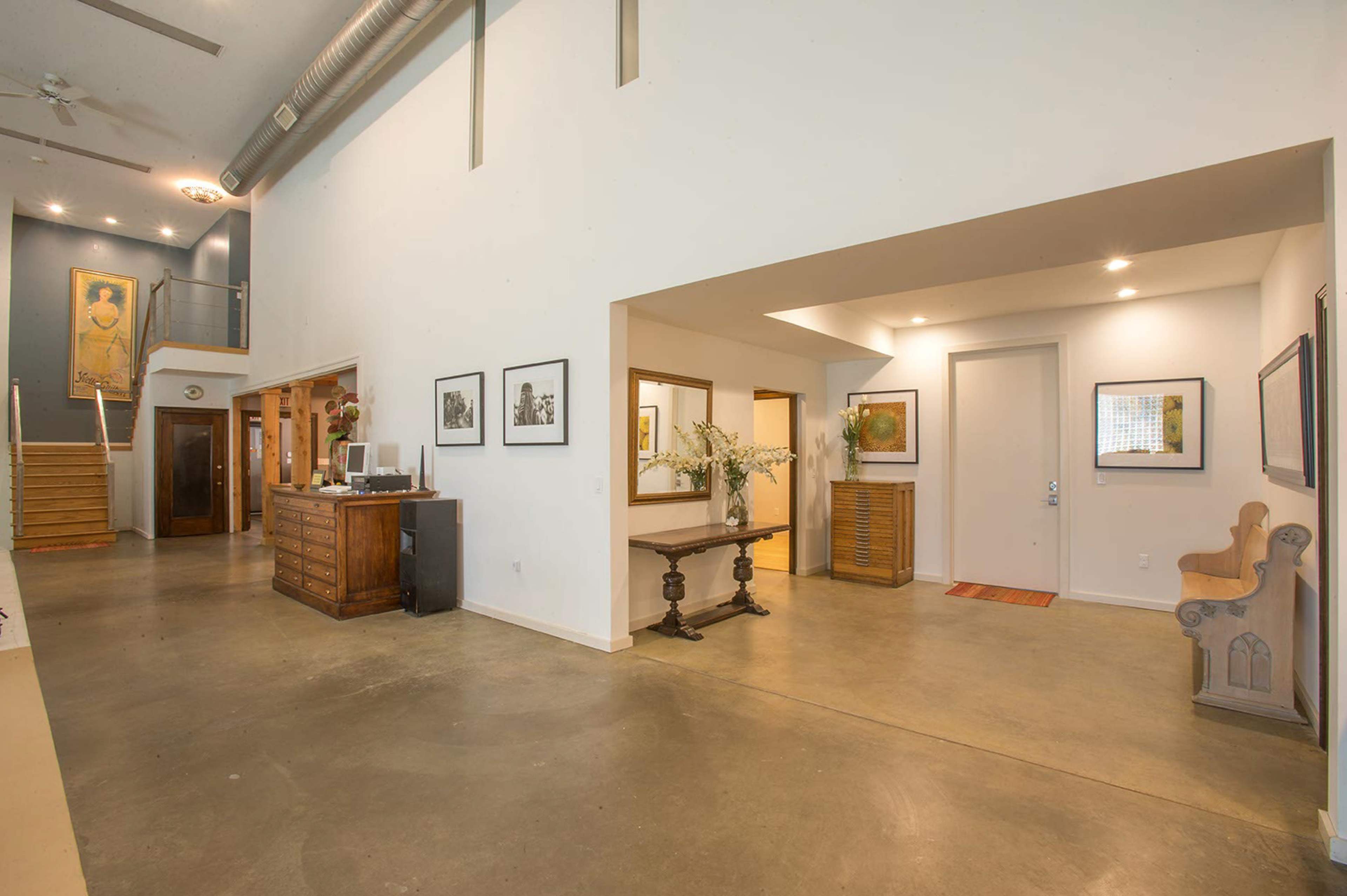A spacious entryway with a concrete floor, featuring a wooden dresser, two framed artworks, and a small table with a vase of flowers.