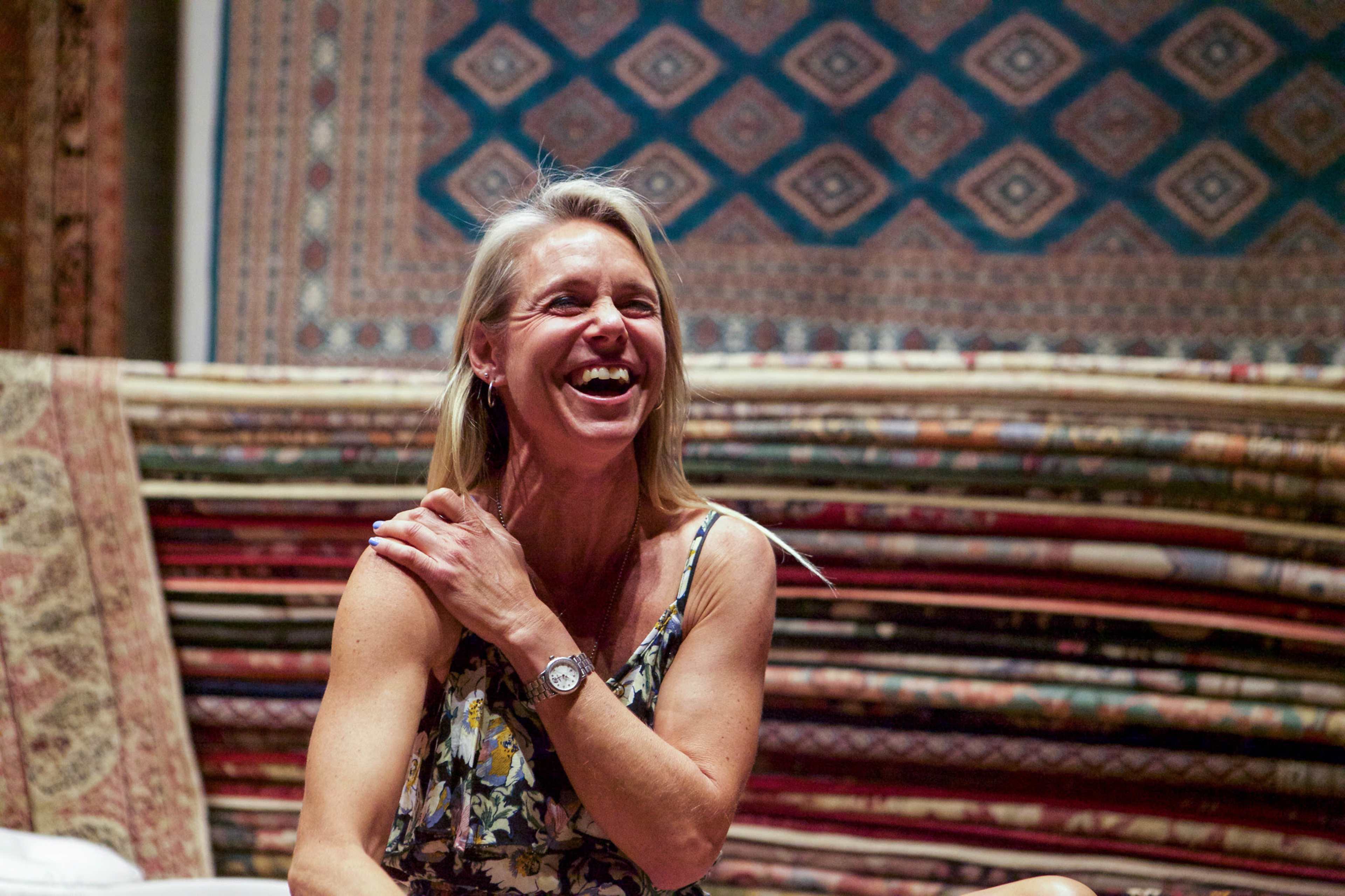 A woman with long blonde hair laughs while seated in front of various colorful rugs.