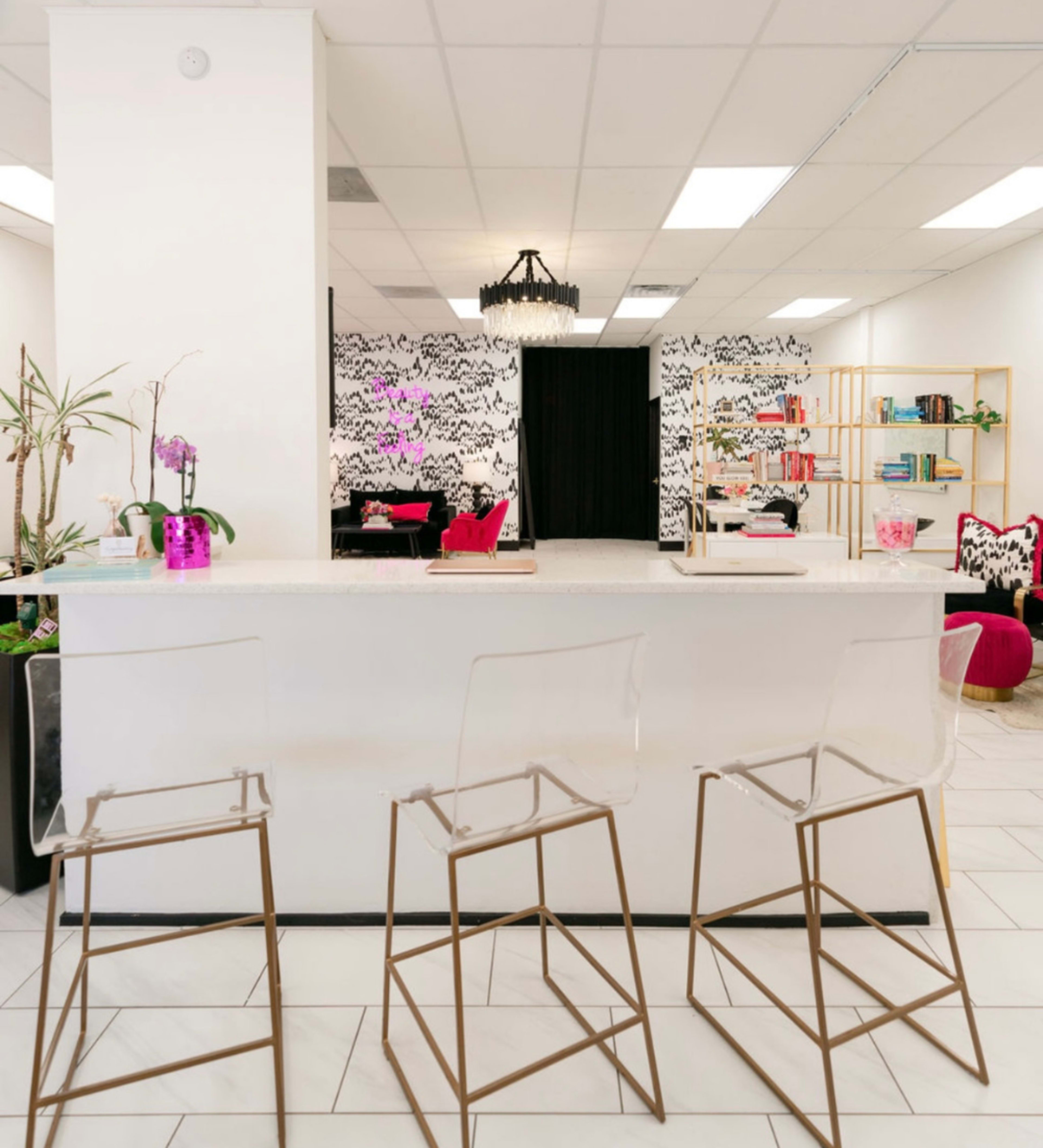 A modern interior features a white bar with three transparent stools, a black and white patterned wall, a chandelier, and shelves filled with books and decorative items.