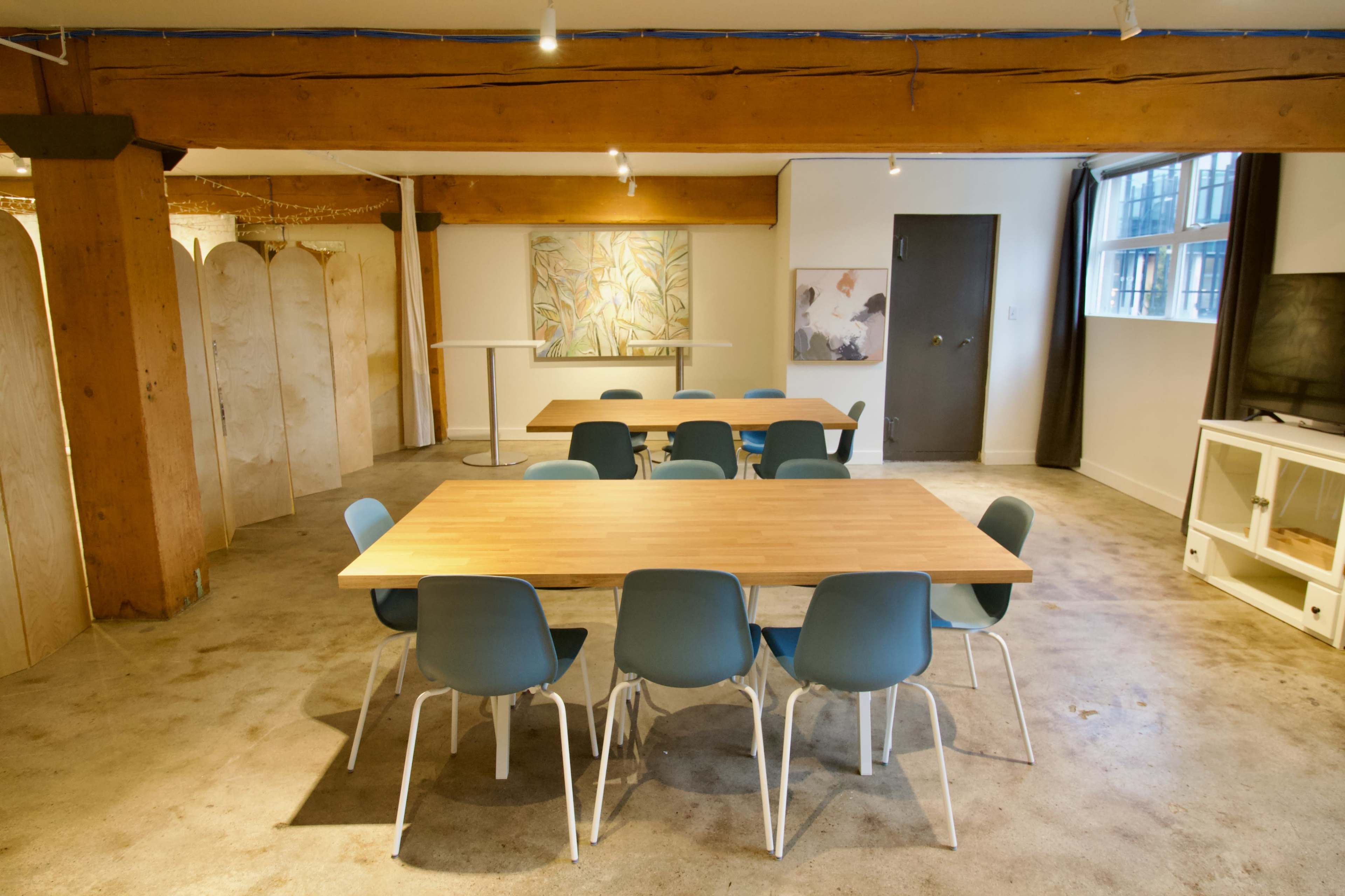 A well-lit interior space features multiple wooden tables surrounded by blue chairs, with artistic decorations visible on the walls.
