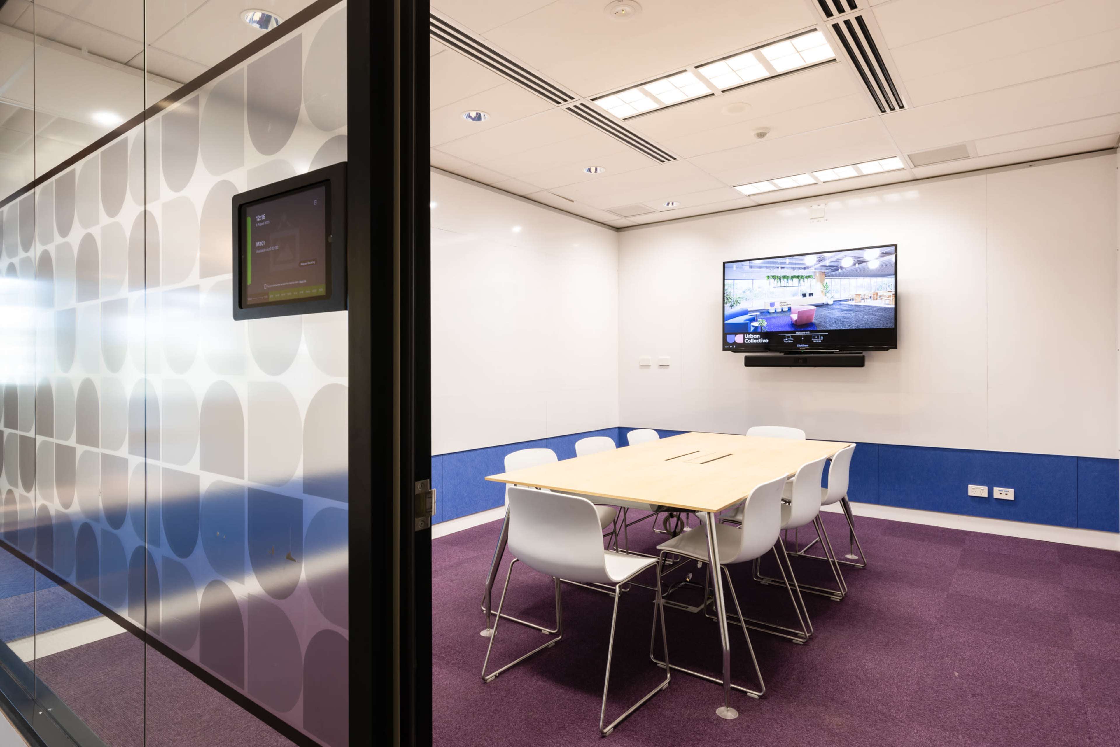 A conference room features a large table surrounded by white chairs, with a television mounted on the wall and a touchscreen control panel nearby.