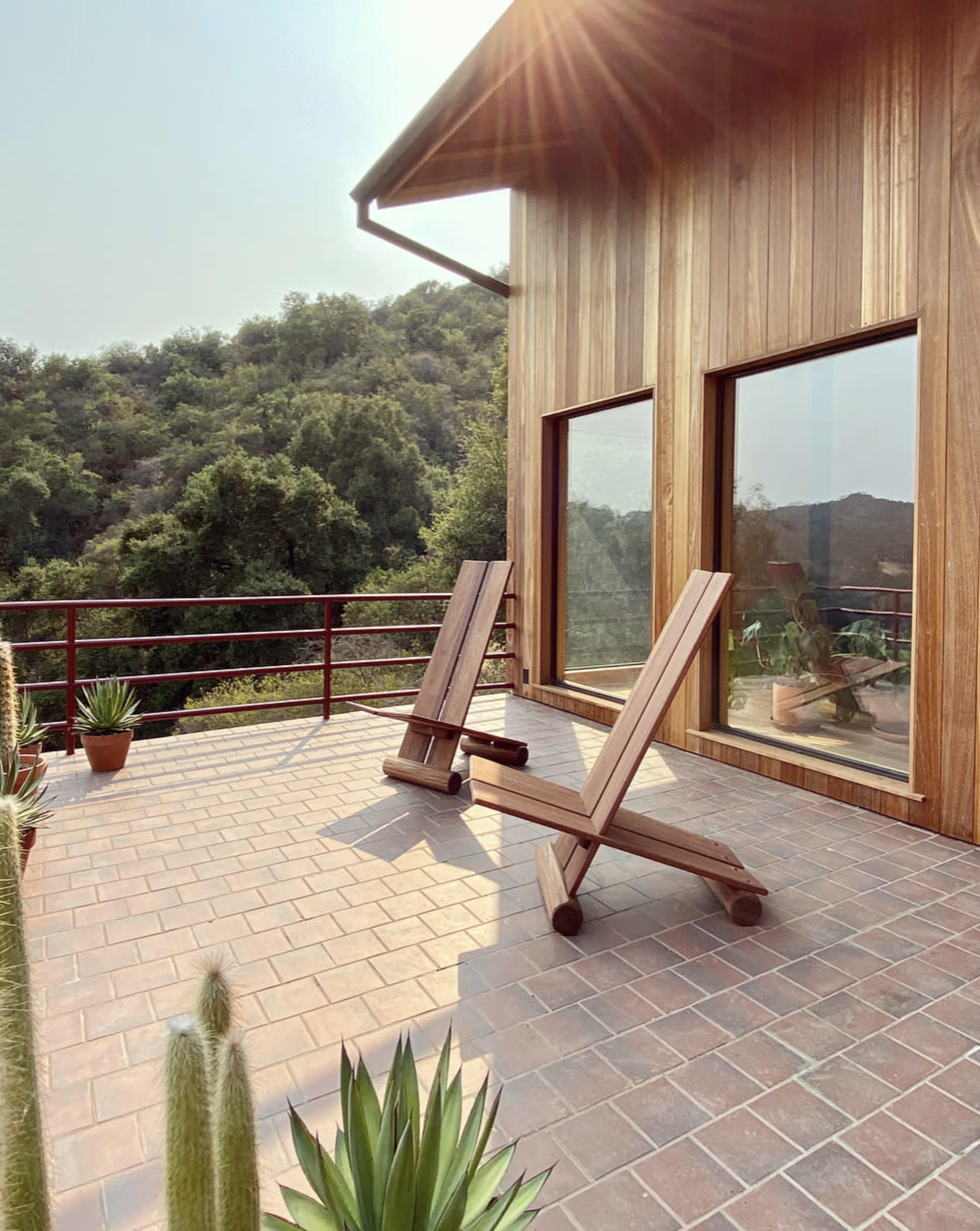 The scene shows a wooden terrace with two sun loungers facing a green hillside, surrounded by potted plants.