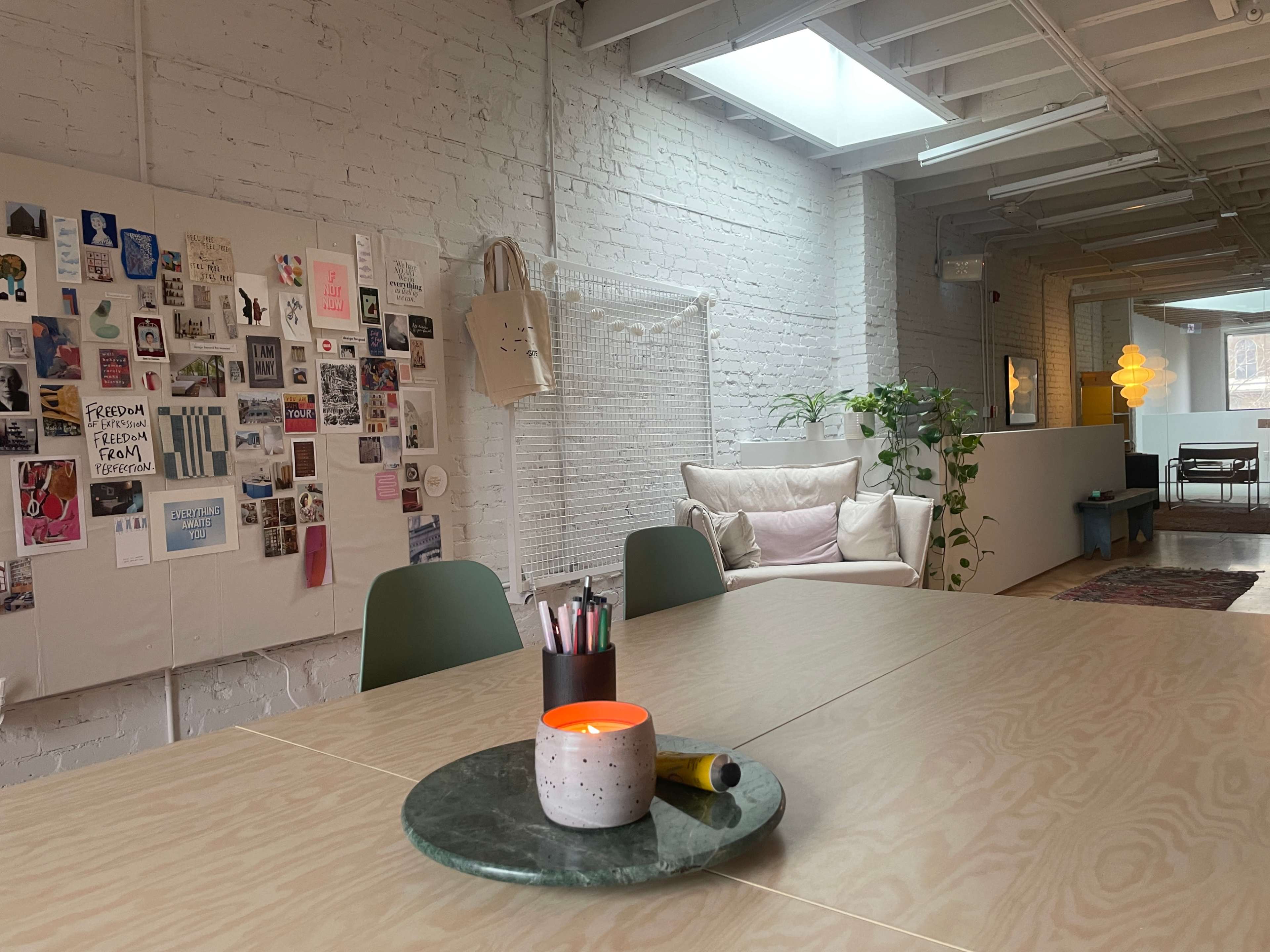 The image shows a modern office space with a large table, various decorations on the wall, and a cozy seating area with a plant.