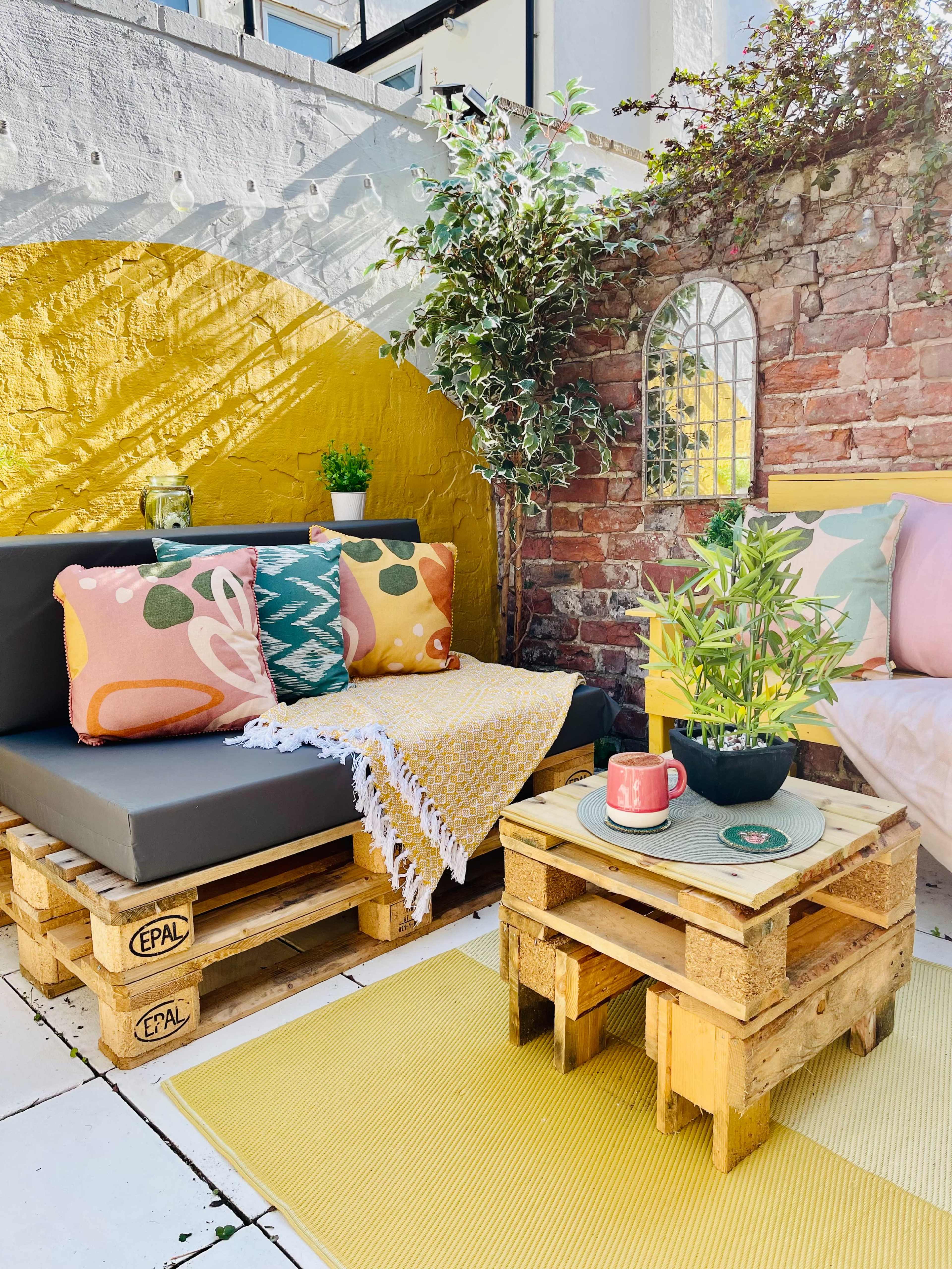 A cozy outdoor seating area with pallet furniture, colorful cushions, a small coffee table, and potted plants against a vibrant yellow and brick wall backdrop.