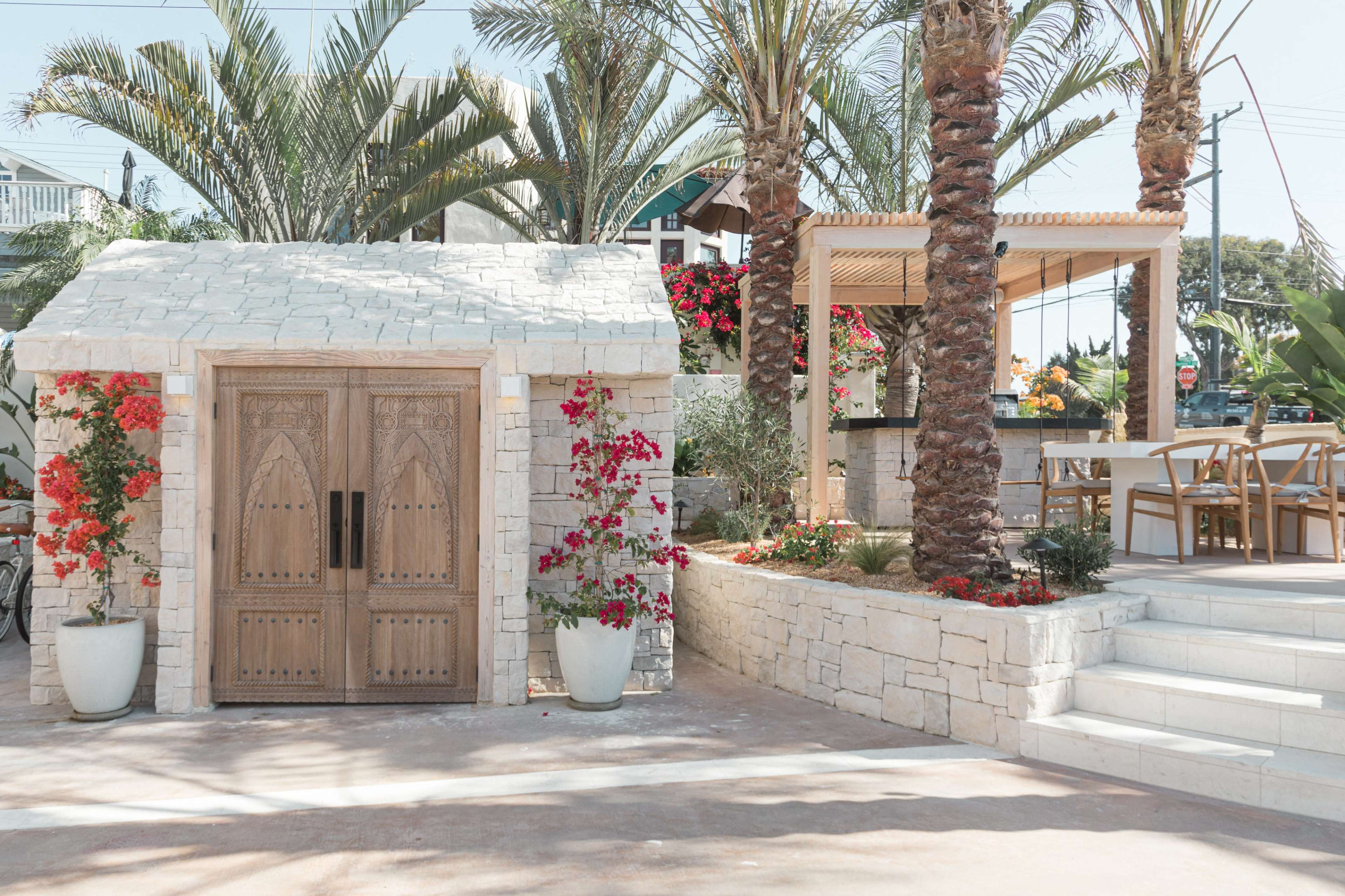 A stone building with ornate wooden doors, surrounded by palm trees and vibrant flowers, next to a shaded seating area with a table and chairs.