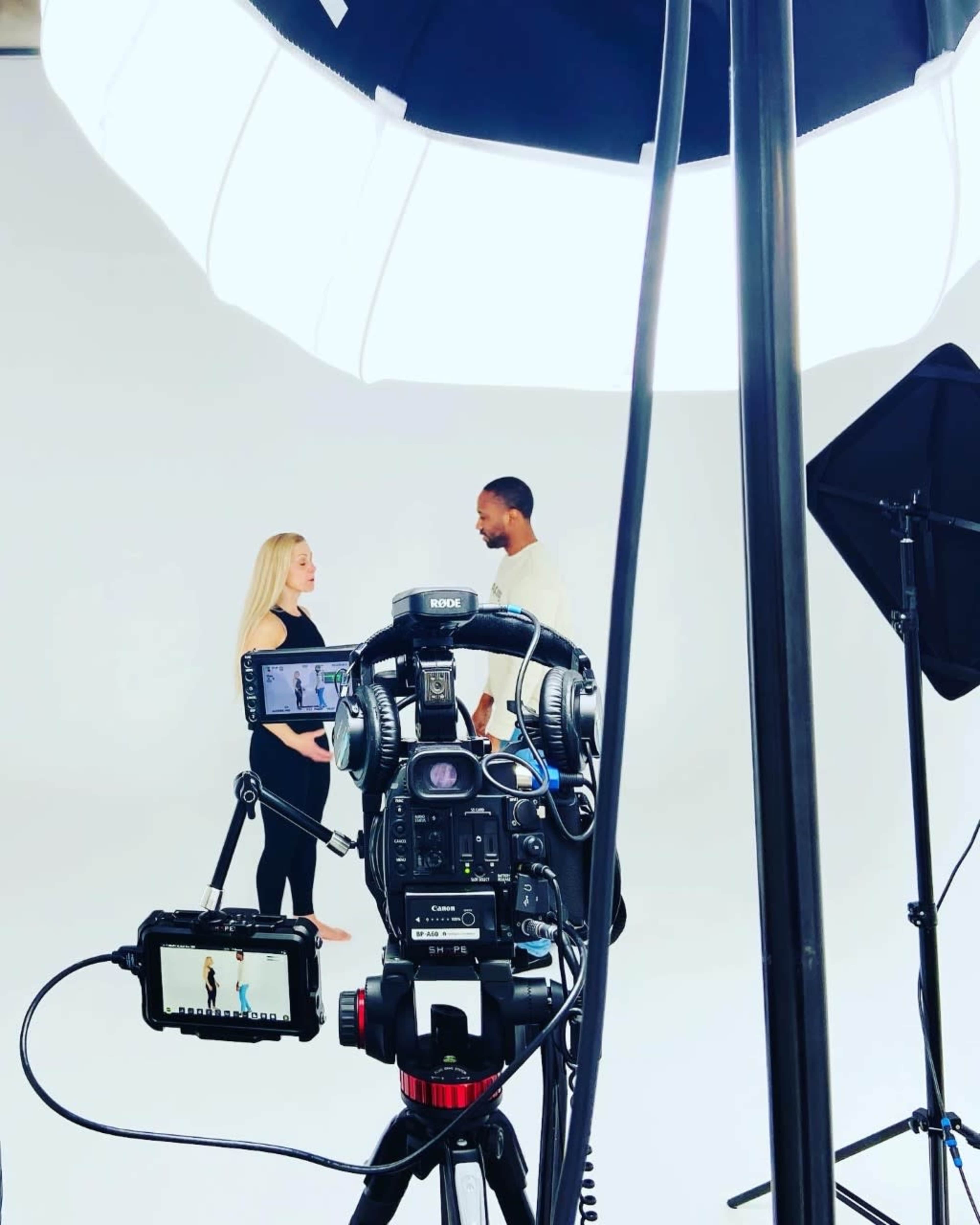 A camera set up on a tripod captures two individuals engaged in conversation in a well-lit studio against a plain white backdrop.
