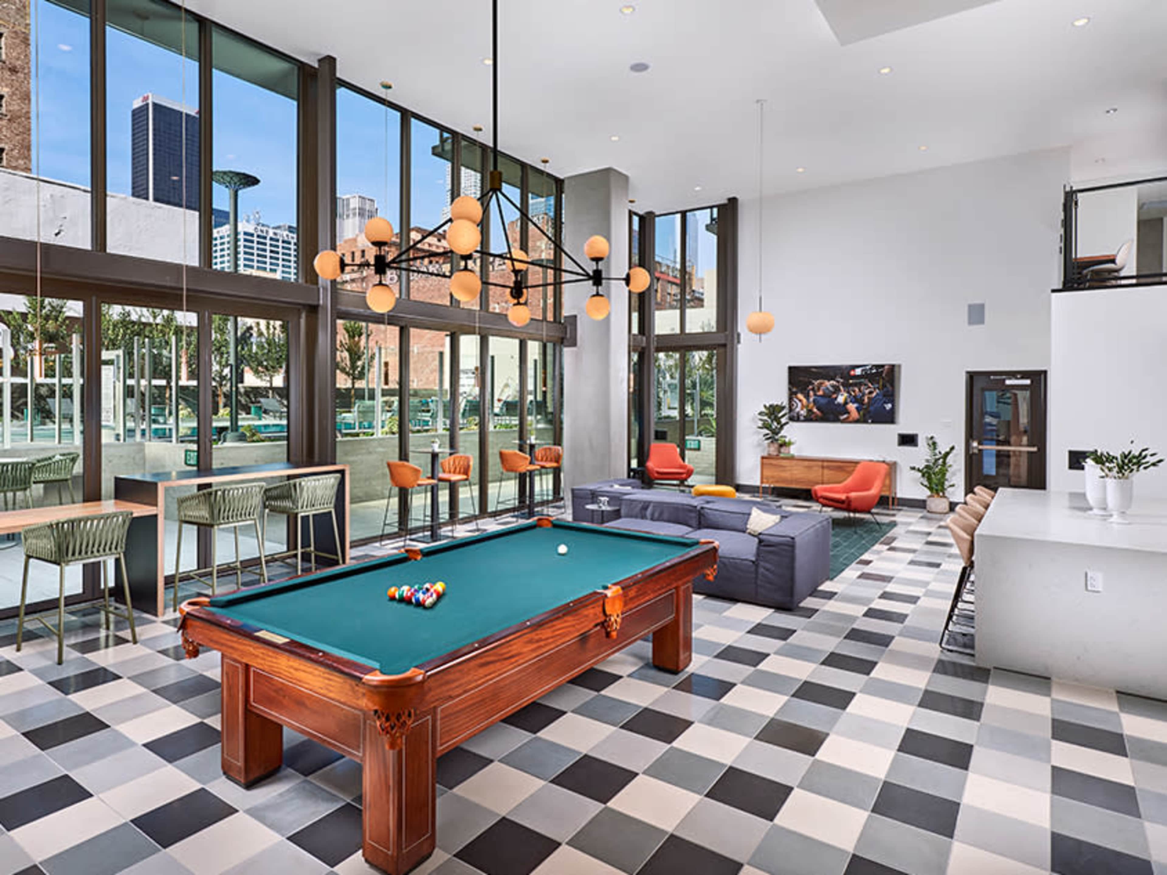 The image shows a modern recreational room featuring a pool table, seating areas, and large windows with urban views.