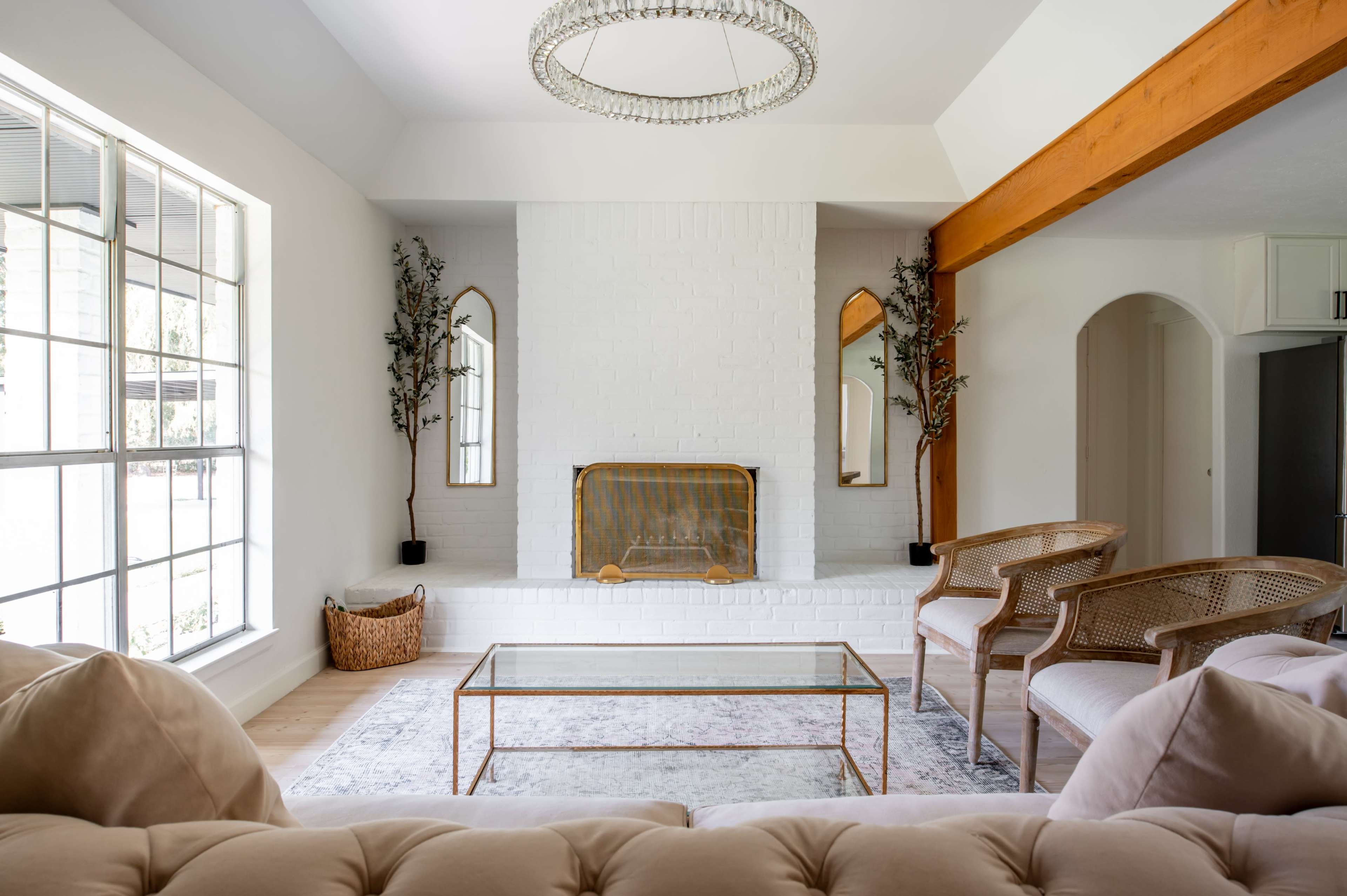 A bright living room features a white brick fireplace, a glass coffee table, and two rattan chairs, framed by large windows and decorative plants.