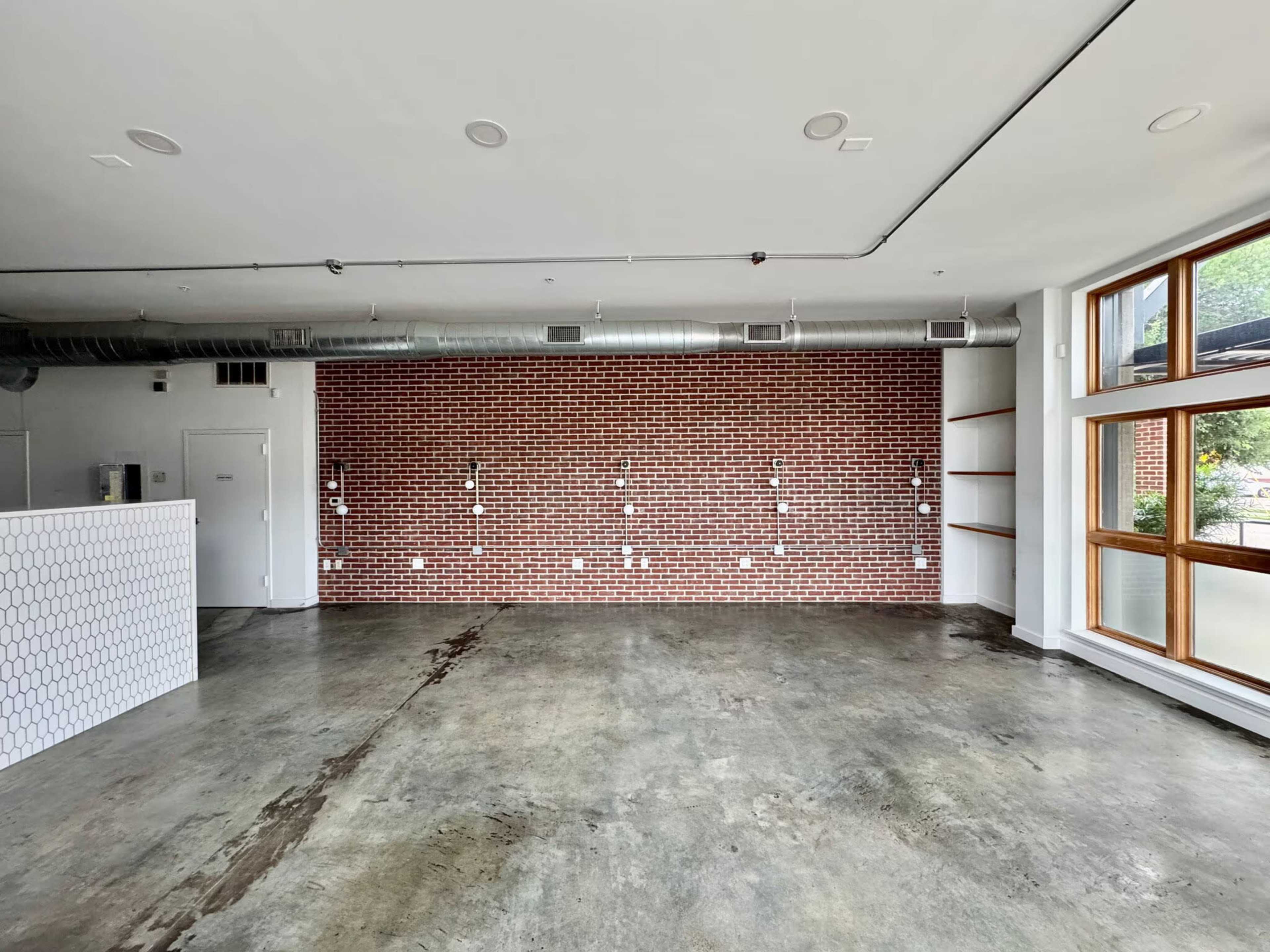 The image shows an empty room with a brick accent wall, a polished concrete floor, and large windows allowing natural light.